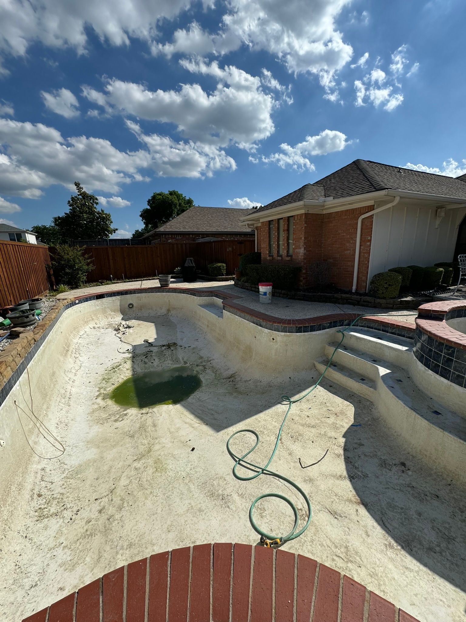 Empty swimming pool with brick coping, house in background, under a blue sky.