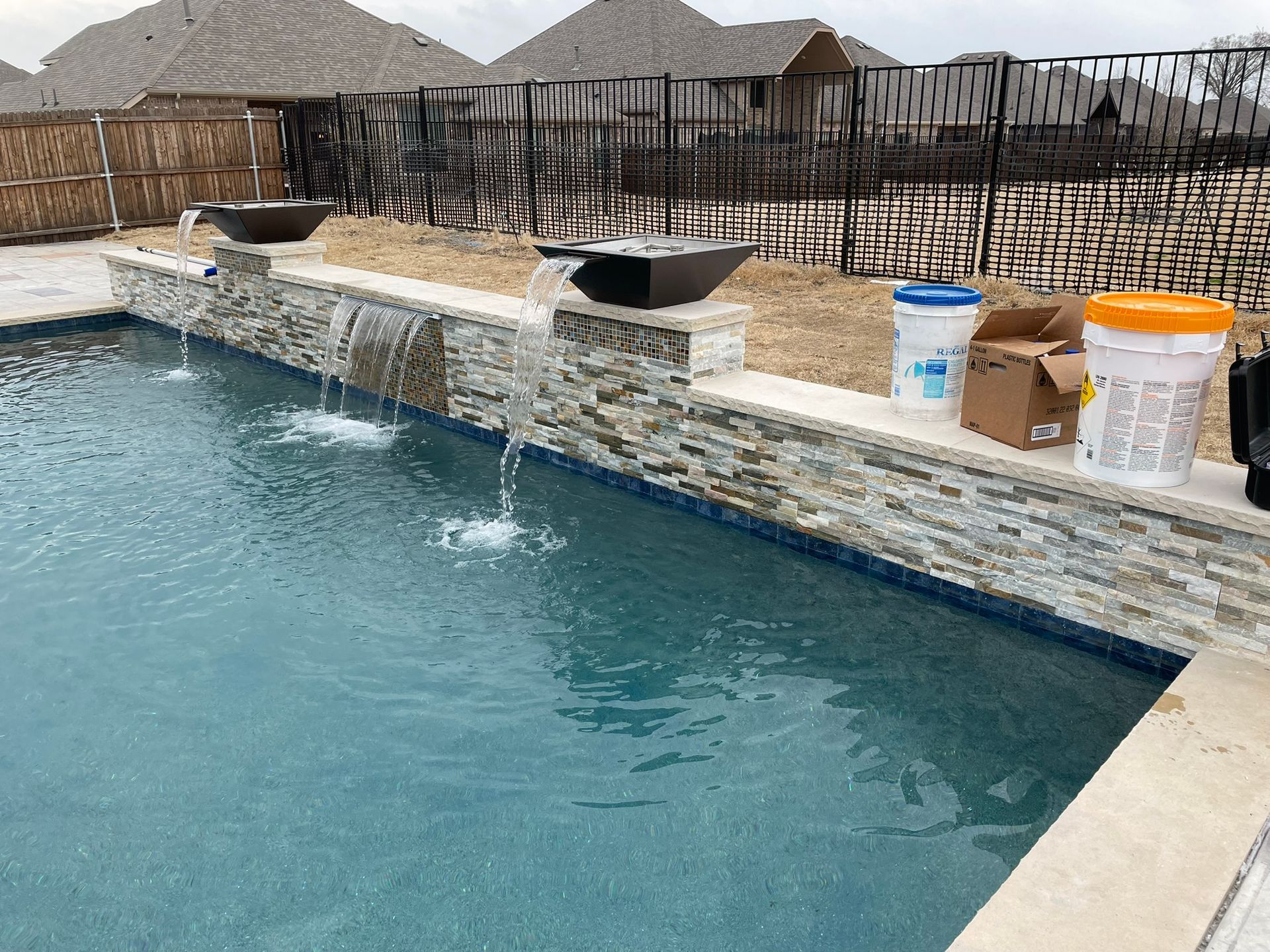 Swimming pool with a stone wall and water fountains; buckets and a box are visible on the wall.