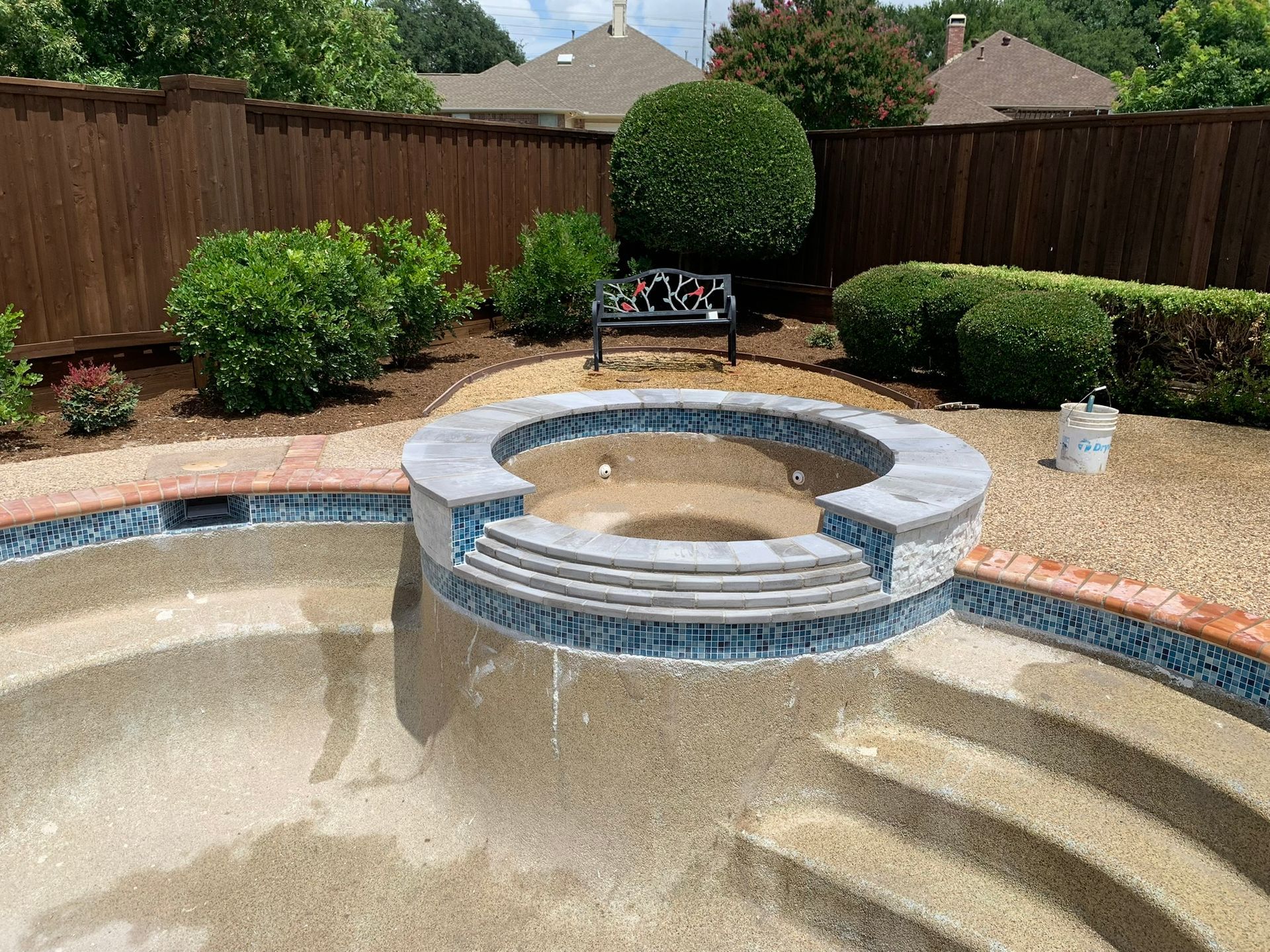 Empty swimming pool with stone steps and blue tile trim, surrounded by landscaping and a wooden fence.