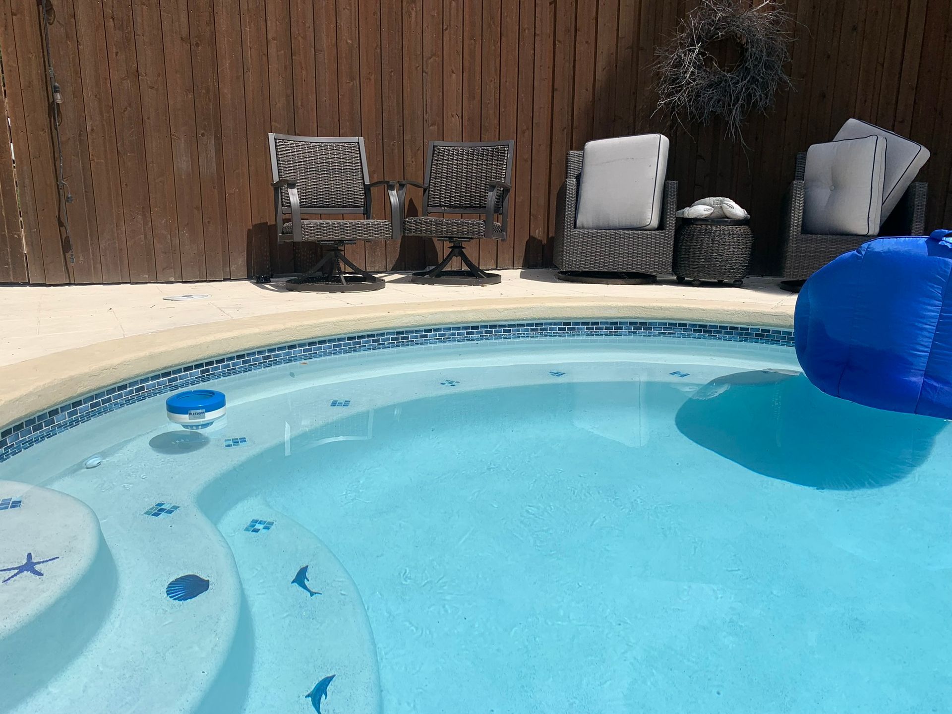 A pool with blue water and steps, surrounded by patio furniture and a wooden fence.