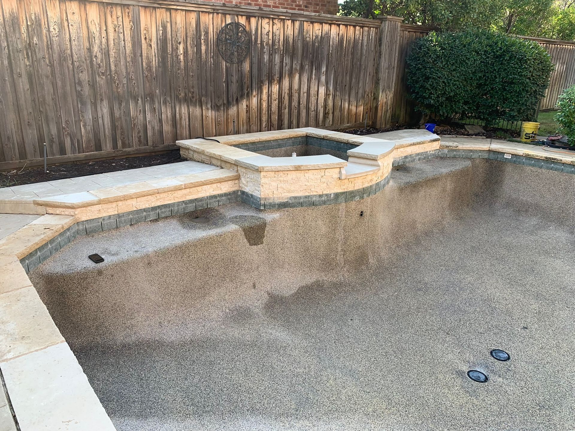 Empty swimming pool with stone coping and a hot tub. Wooden fence in background.