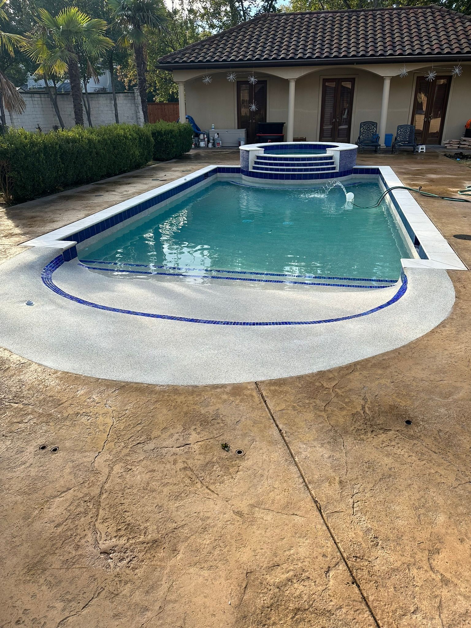 Pool with blue tile trim and light blue water, surrounded by concrete and a small building in the background.