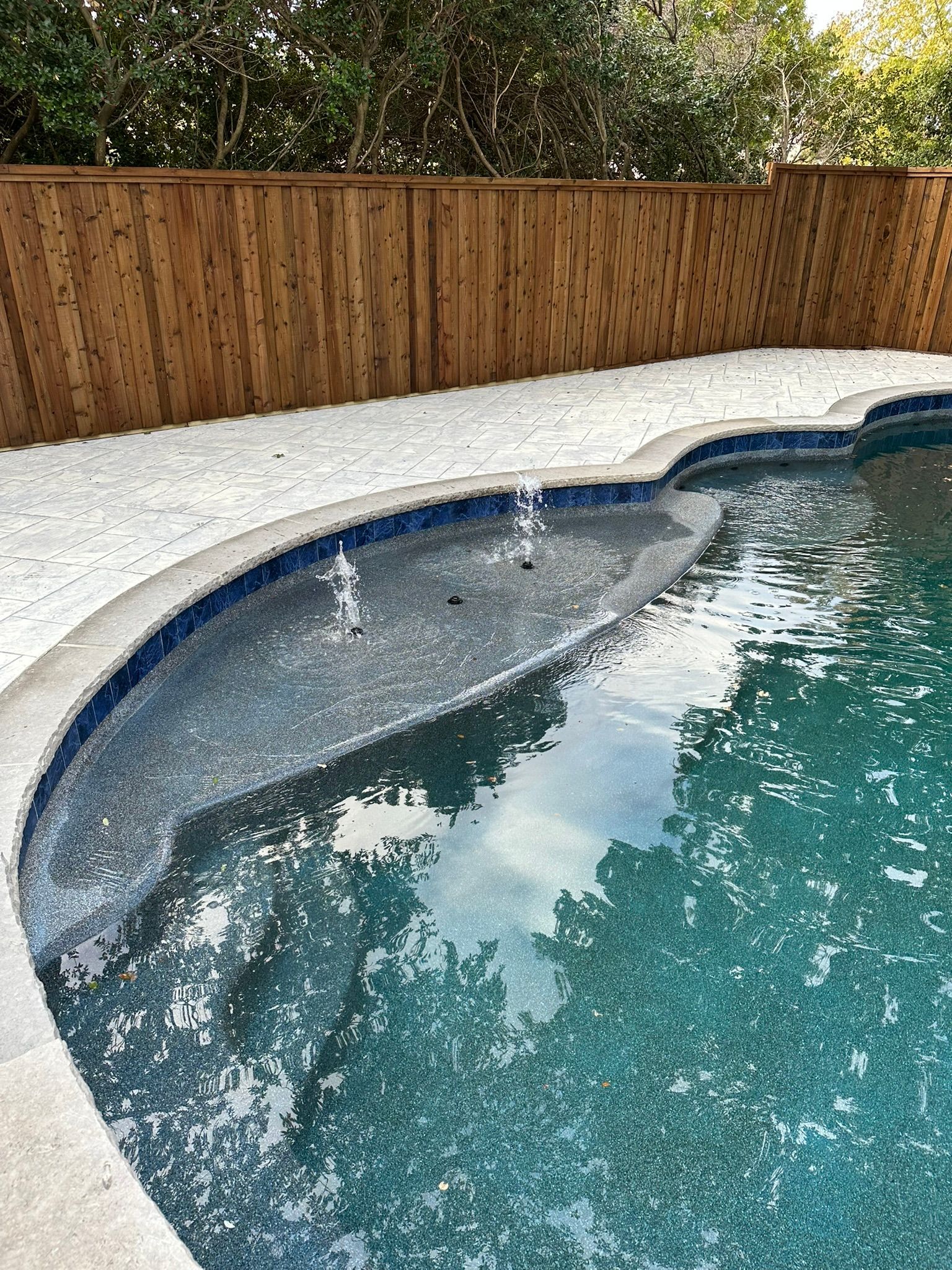 Pool with dark blue mosaic tile, concrete edge, water, white gravel, and wooden fence.