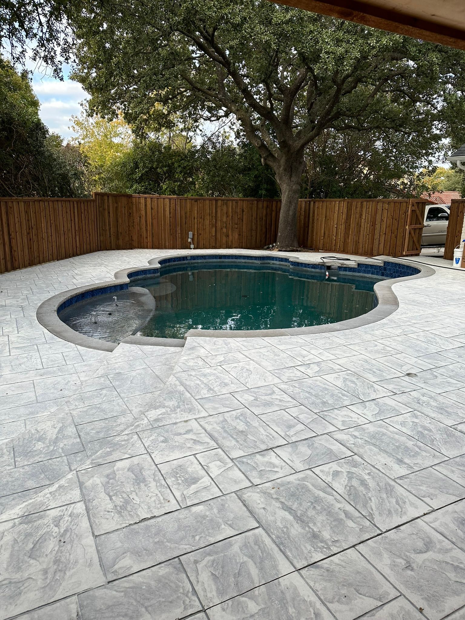 Pool with blue water surrounded by stone patio, wooden fence, and large tree.