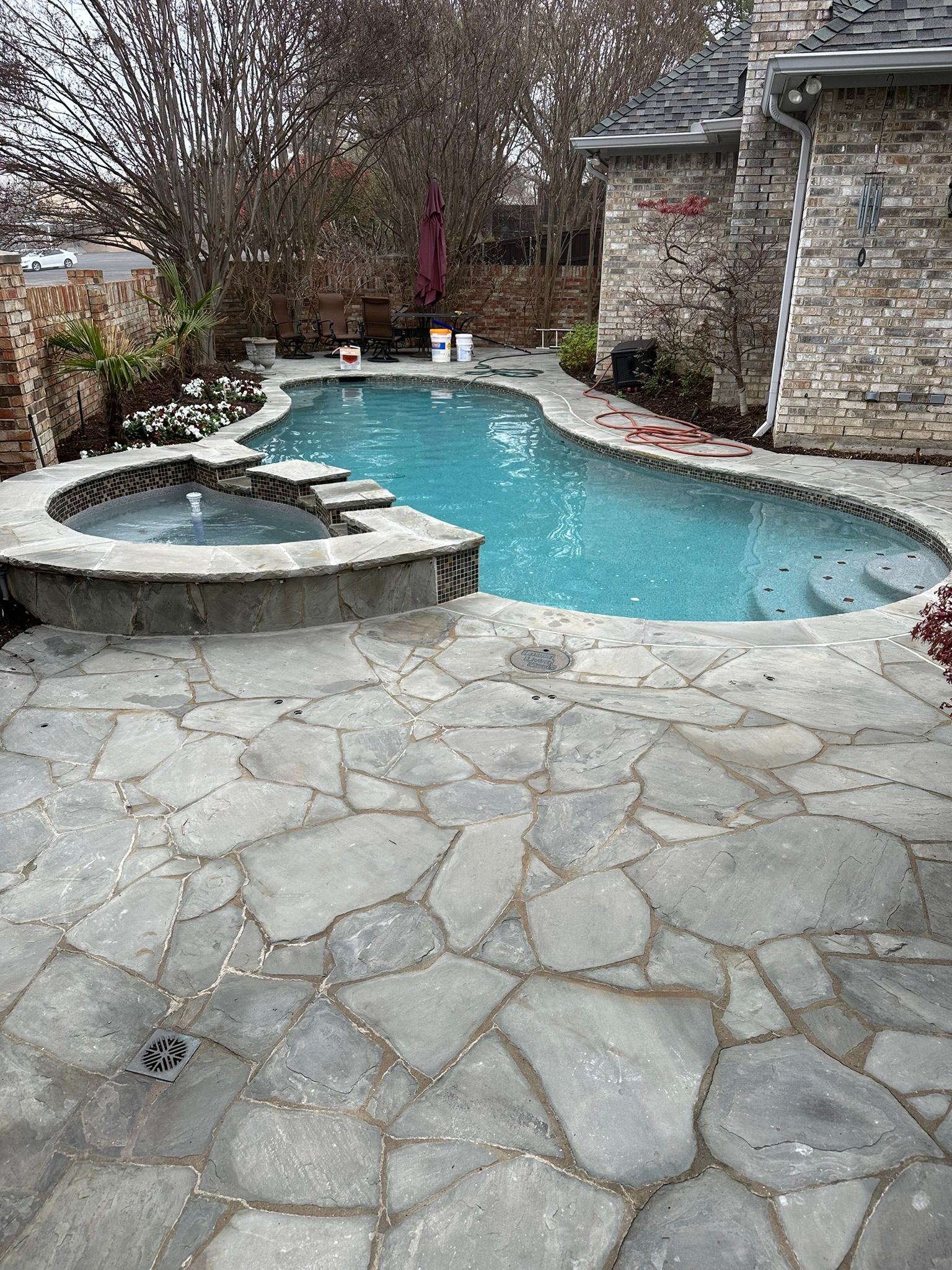 A swimming pool and spa surrounded by stone patio, next to a stone house. Blue water, outdoor setting.