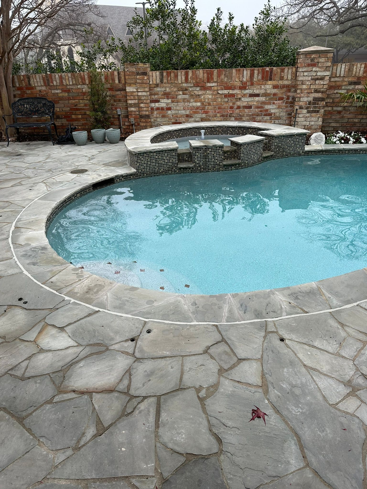 Swimming pool with stone patio, small hot tub, brick wall, and lush greenery.
