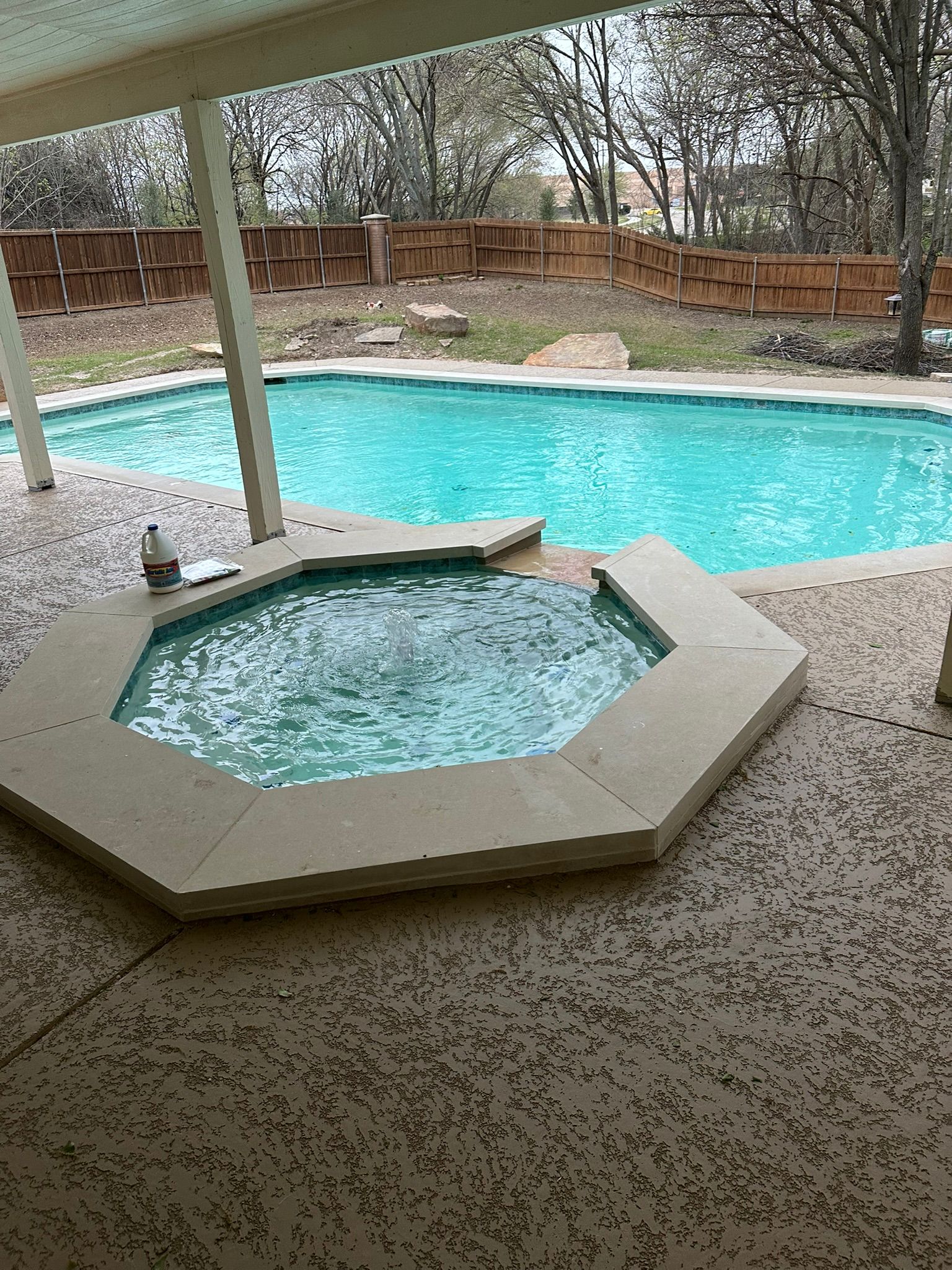 A backyard with a pool and hot tub, tan patio, and wooden fence.