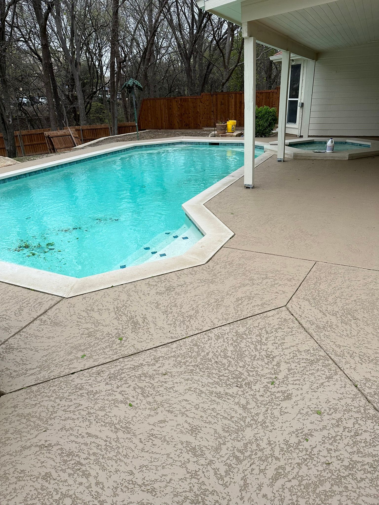 Pool with turquoise water surrounded by textured tan concrete patio and a wooden fence in the background.