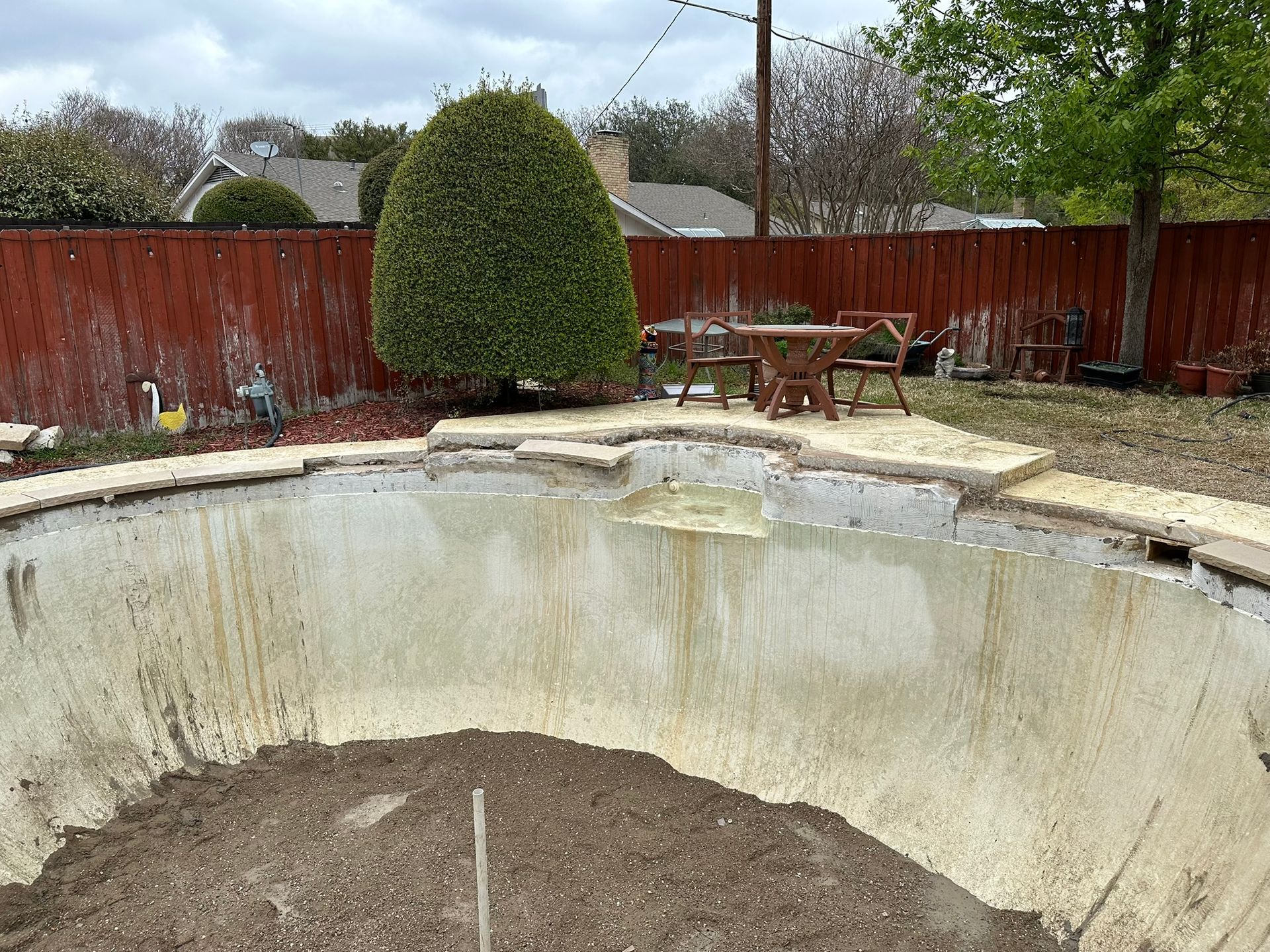 Empty swimming pool in a backyard with a wooden fence and a table and chairs.