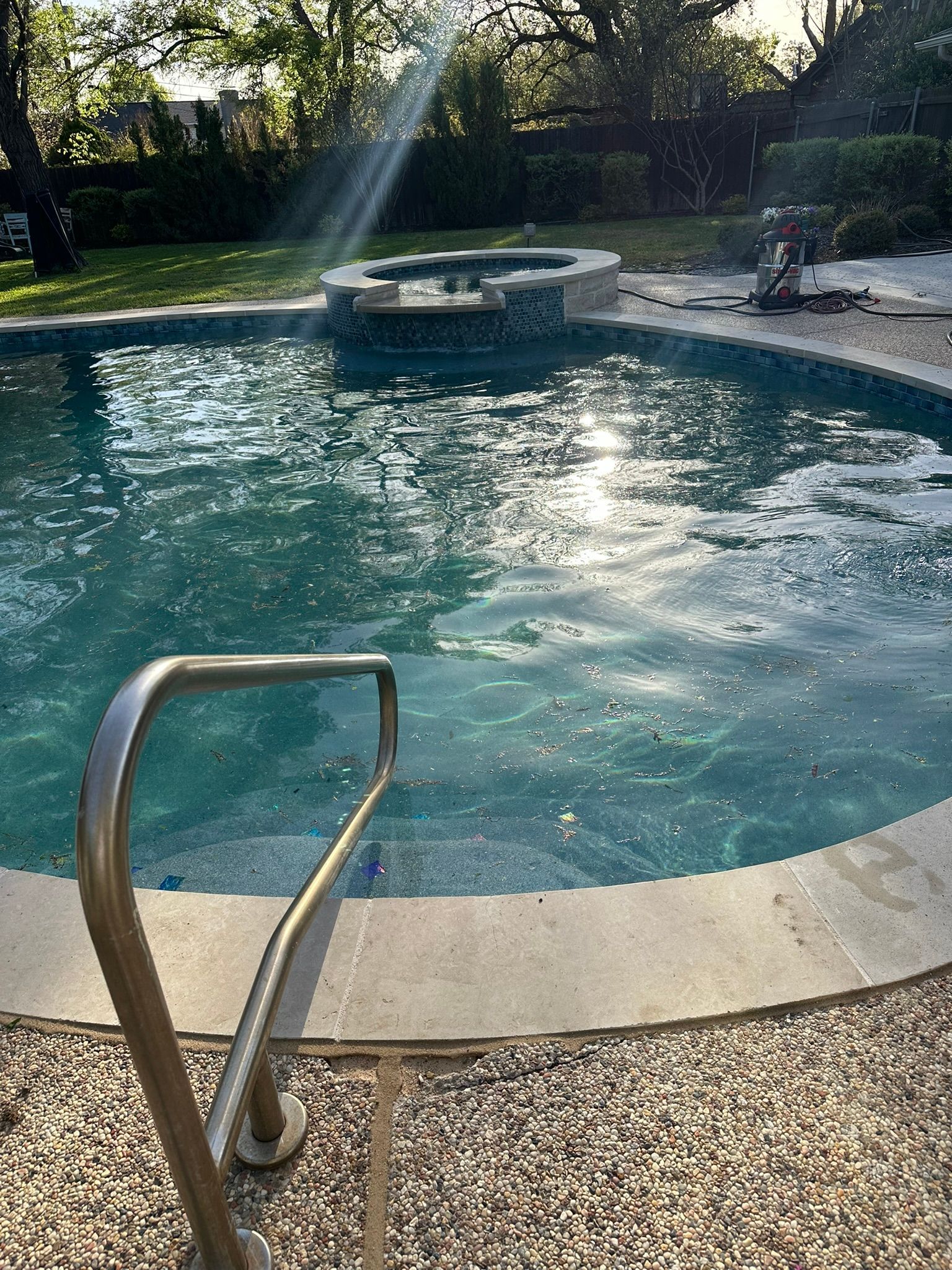 Pool with spa, surrounded by stone and a lawn. A handrail is in the foreground. Sunlight glints on the water.