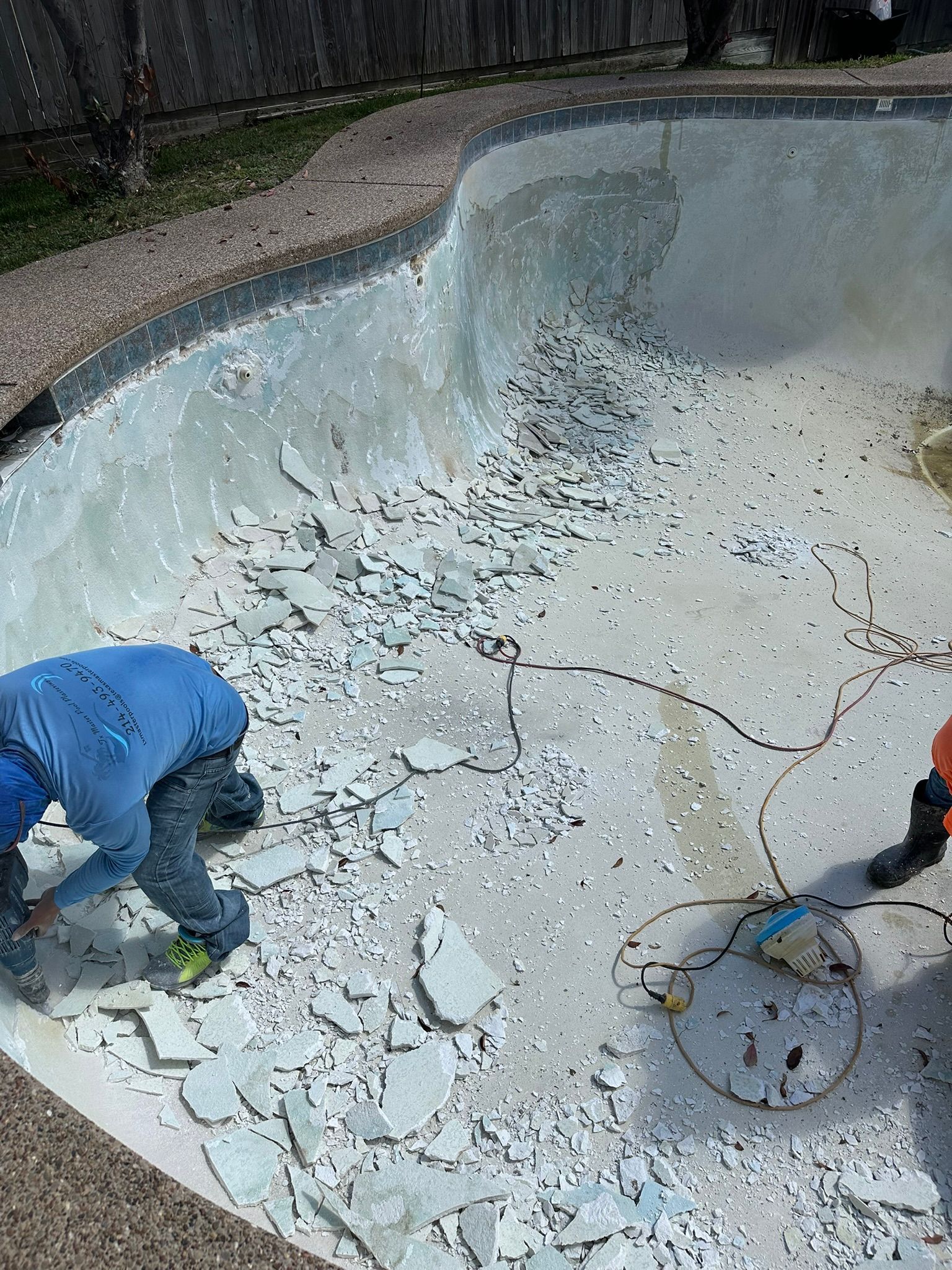Two workers demolish a concrete pool. Broken pieces cover the bottom.