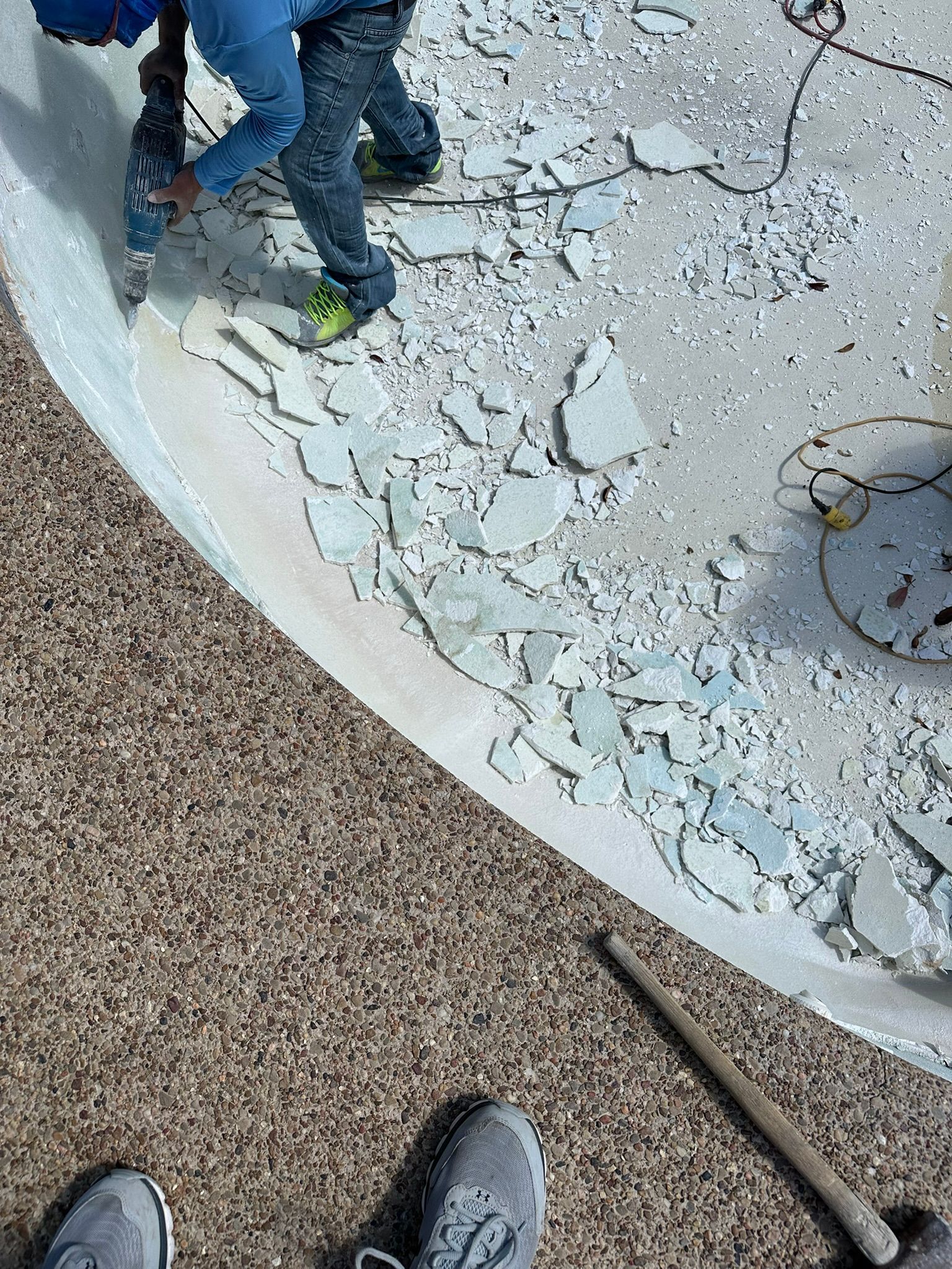 Man using a power tool to remove tiles from a pool. Concrete and scattered debris on the edge.