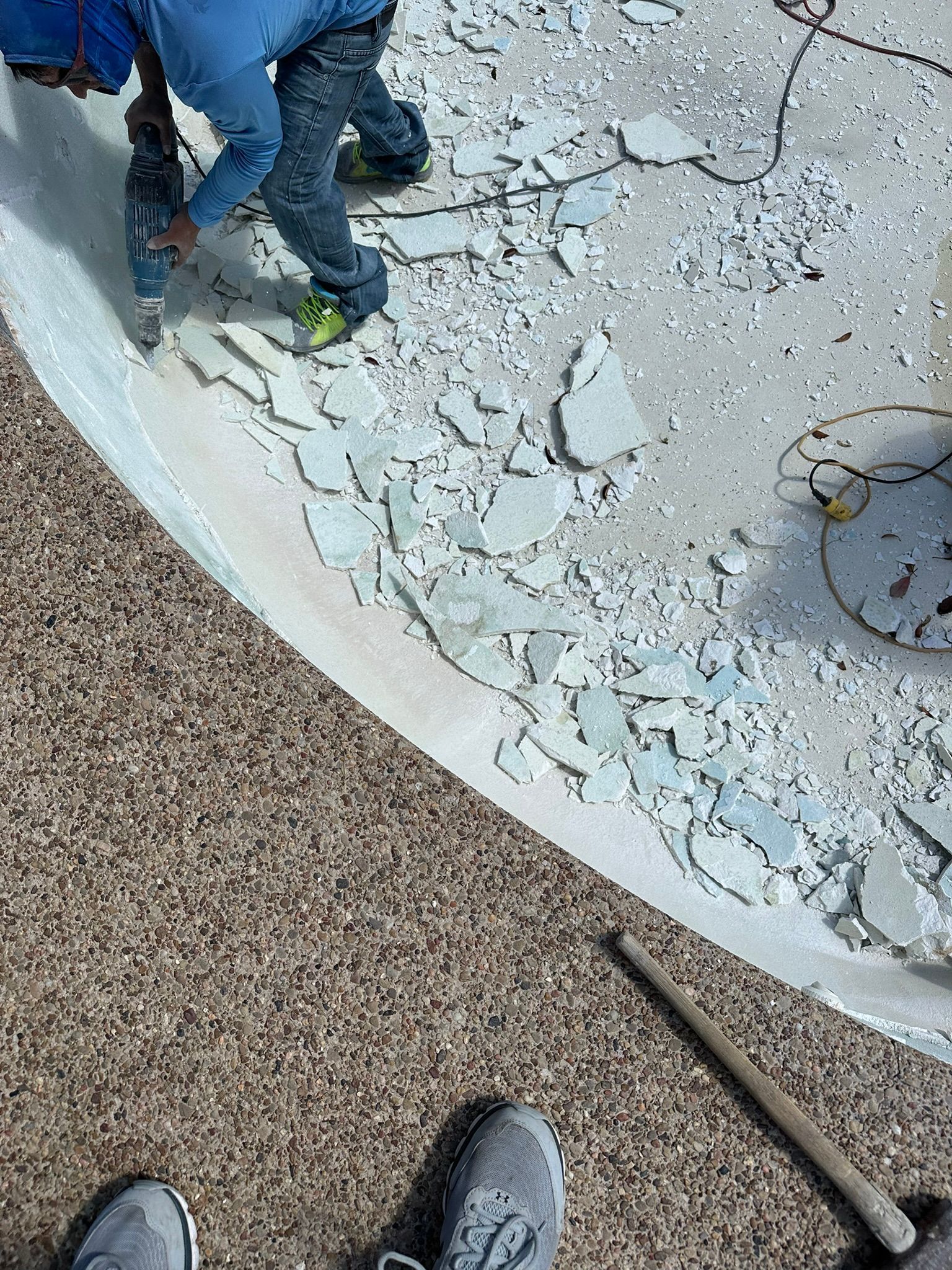 Person using a jackhammer to remove tile from a pool. Brown pebble surrounding the pool.