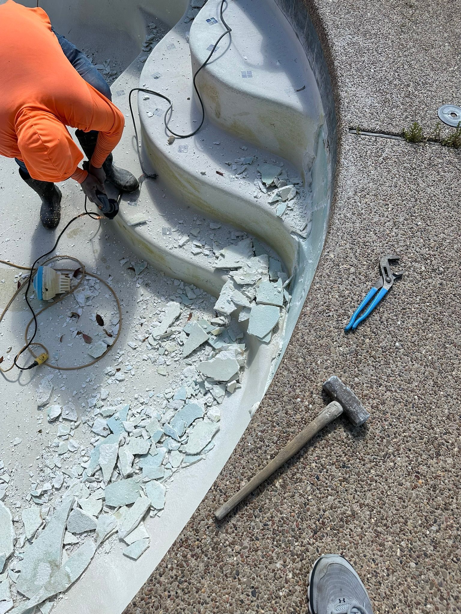Person in orange gloves and sleeves chipping away tiles from a pool with a hammer.