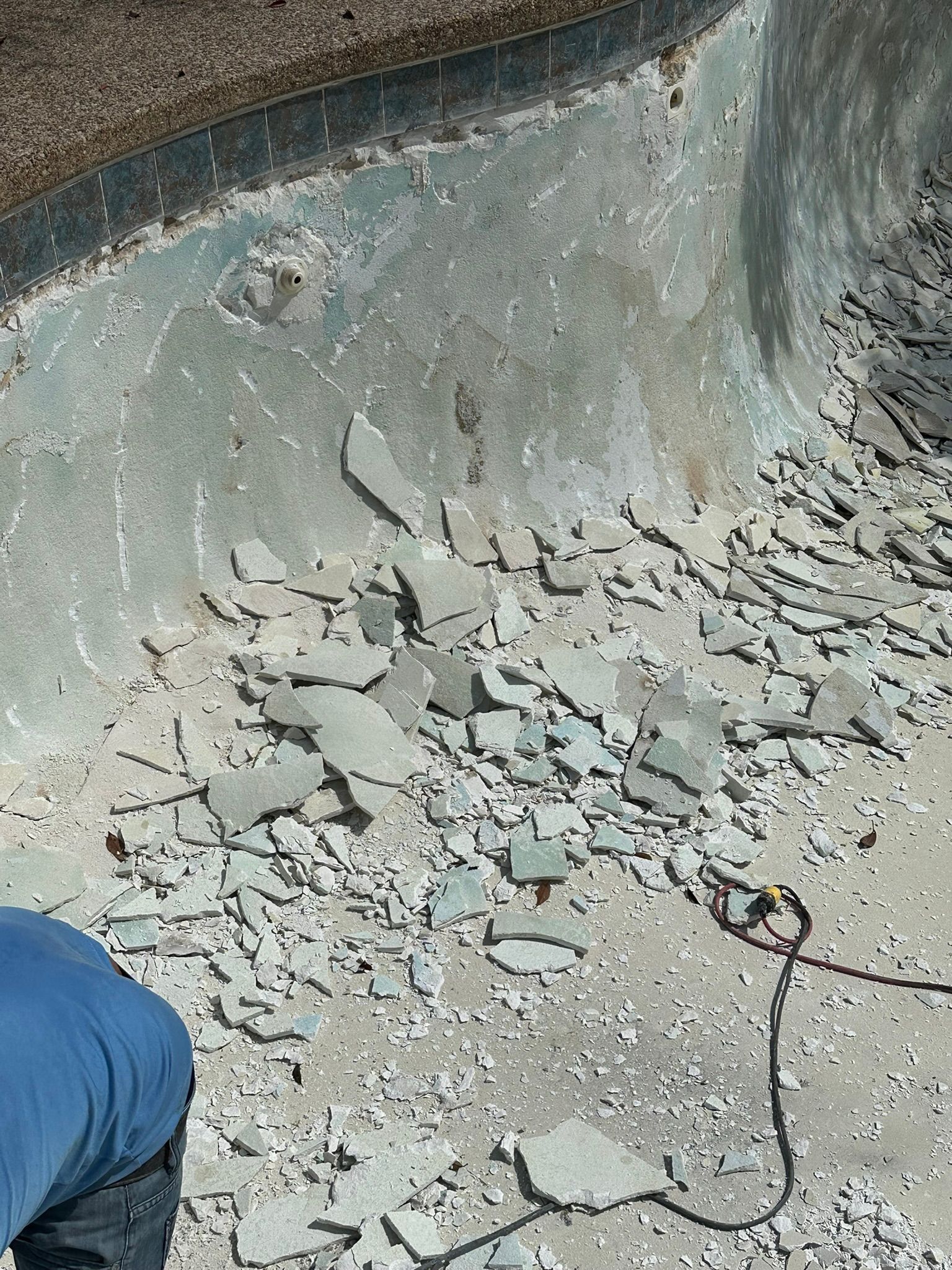 Pool interior with chipped surface and debris. Person in blue shirt working. Concrete setting.