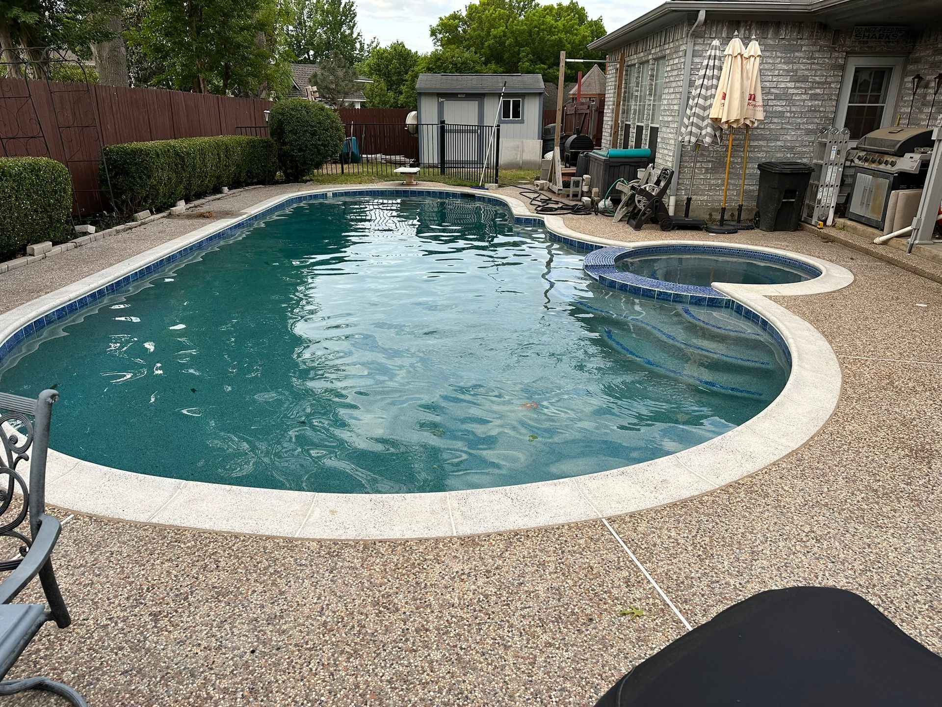 Pool with blue water and a tiled edge, surrounded by a pebbled patio. A hot tub is in the corner.