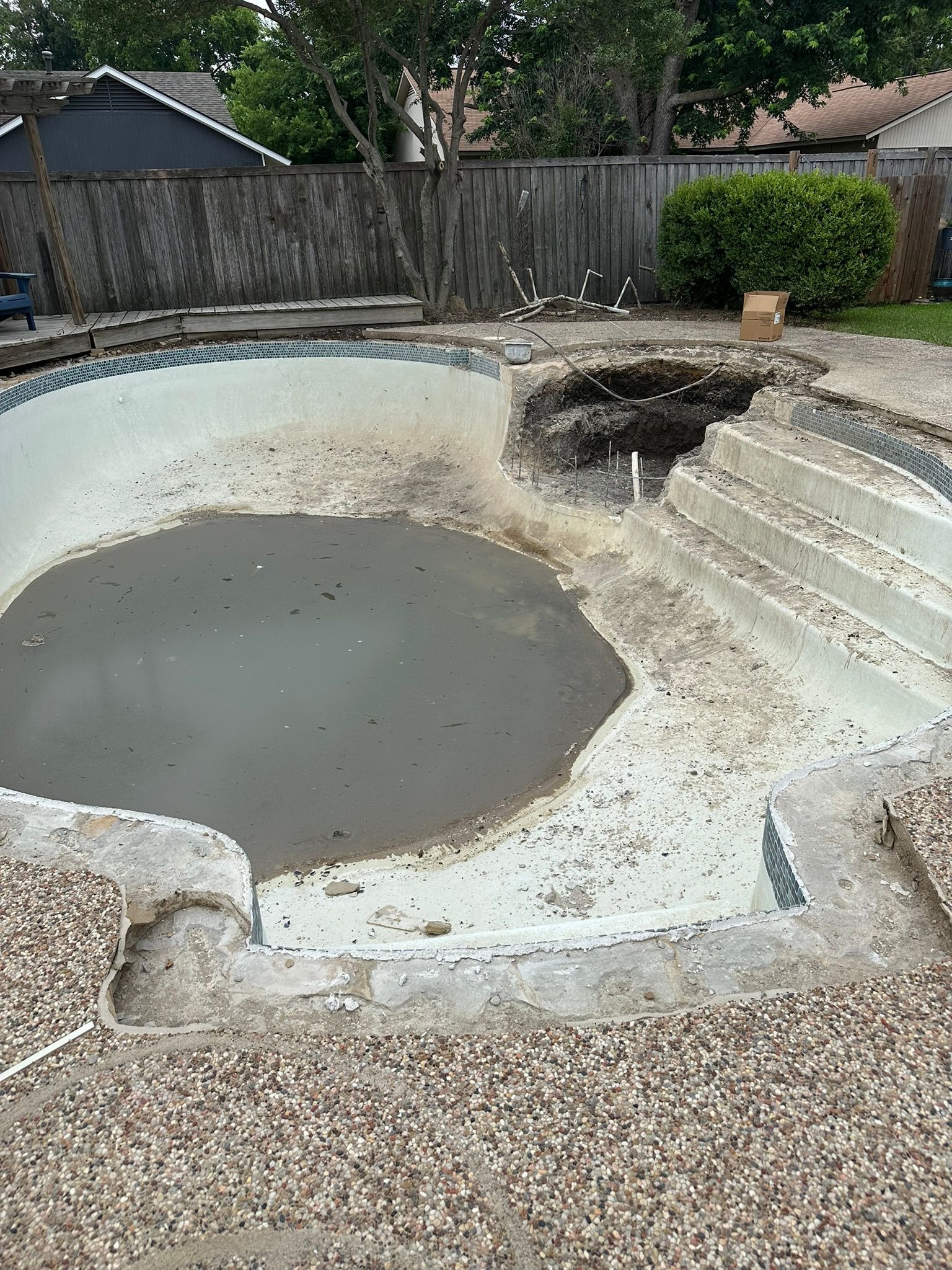 An empty, dirty swimming pool with exposed concrete, steps, and a partially removed deck.