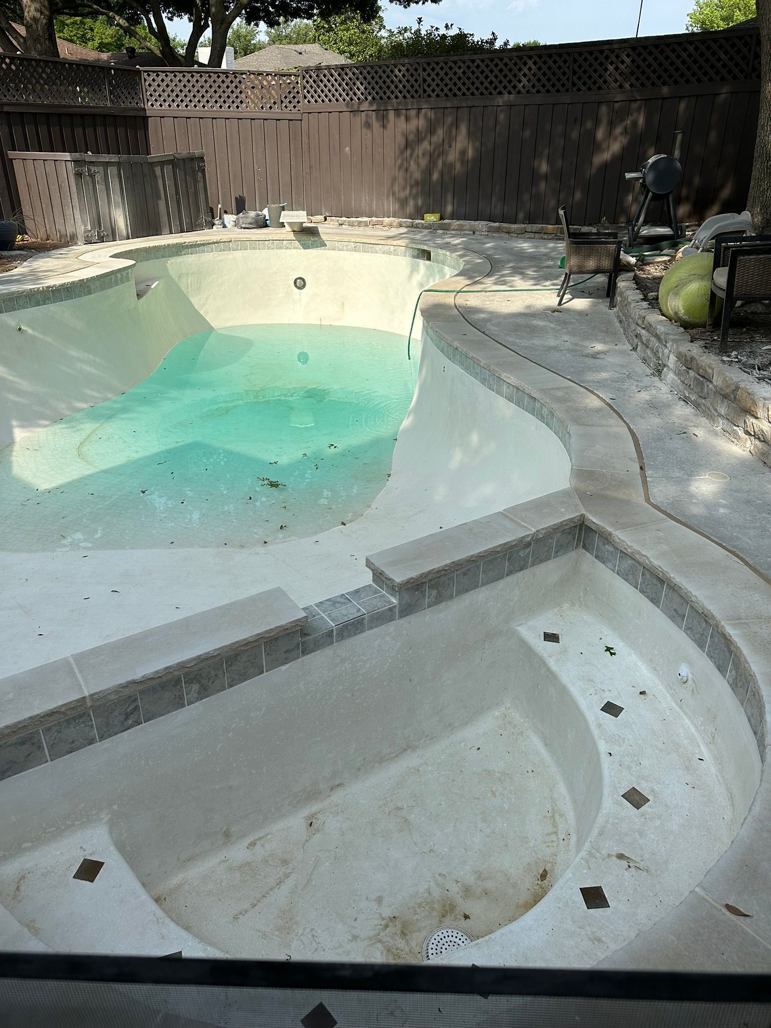 Empty swimming pool with curved steps and turquoise water, surrounded by concrete and a brown fence.