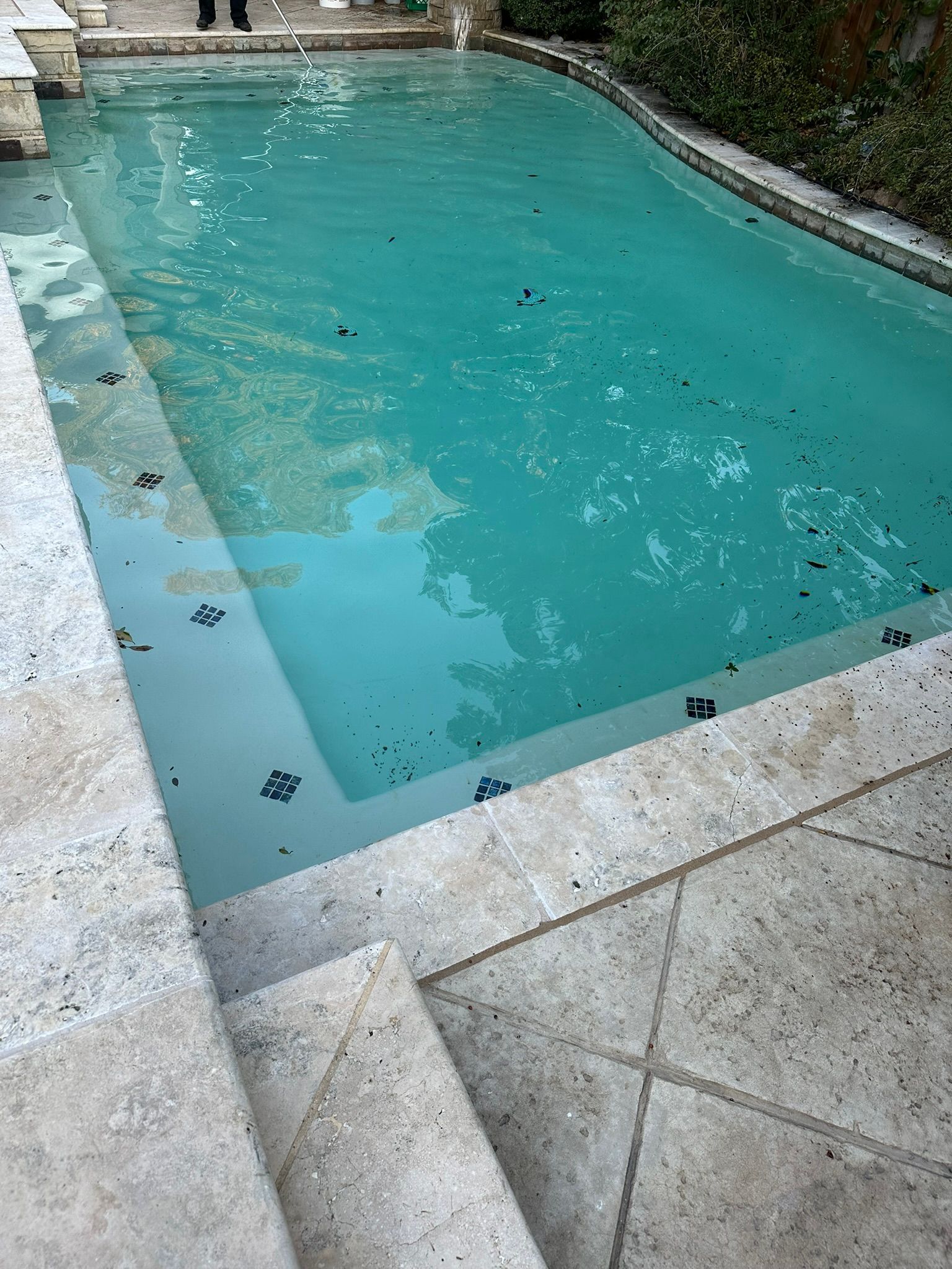 A turquoise swimming pool with stone steps and tiles; debris floats on the water.