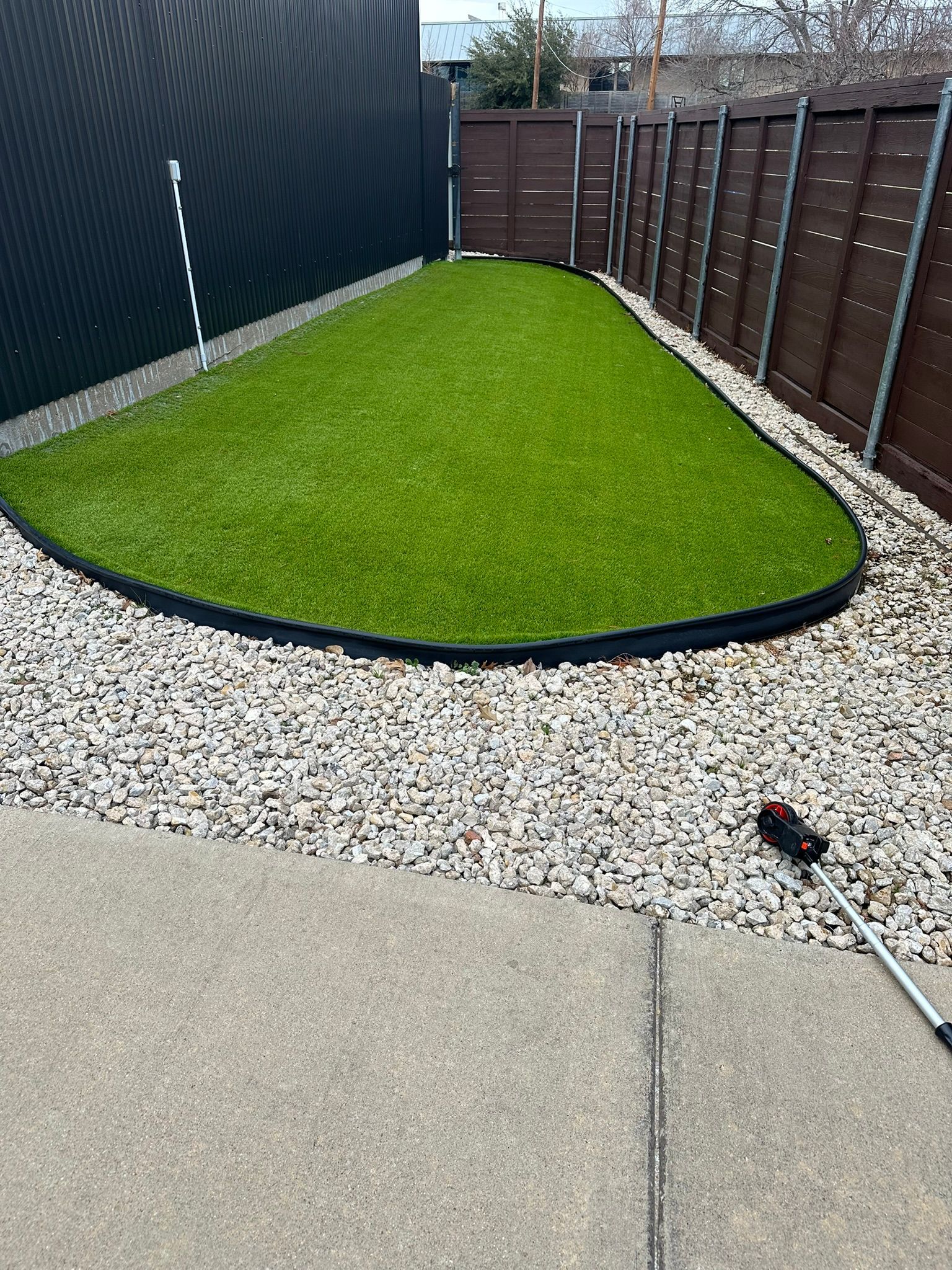 Small artificial turf patch surrounded by gravel and fences. Concrete patio and tool visible.