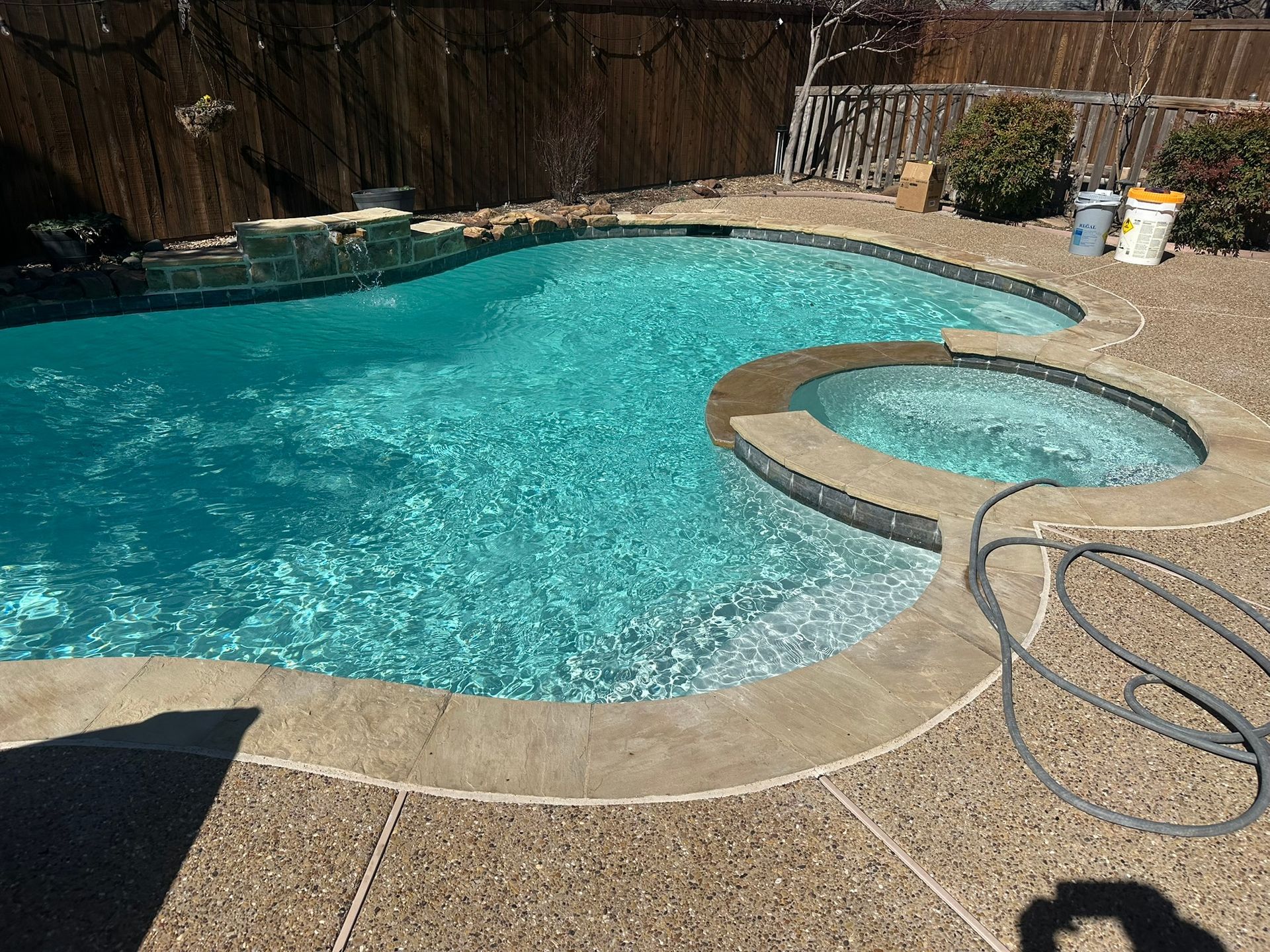 Swimming pool with attached jacuzzi. Turquoise water, tan concrete, brown fence in the background.