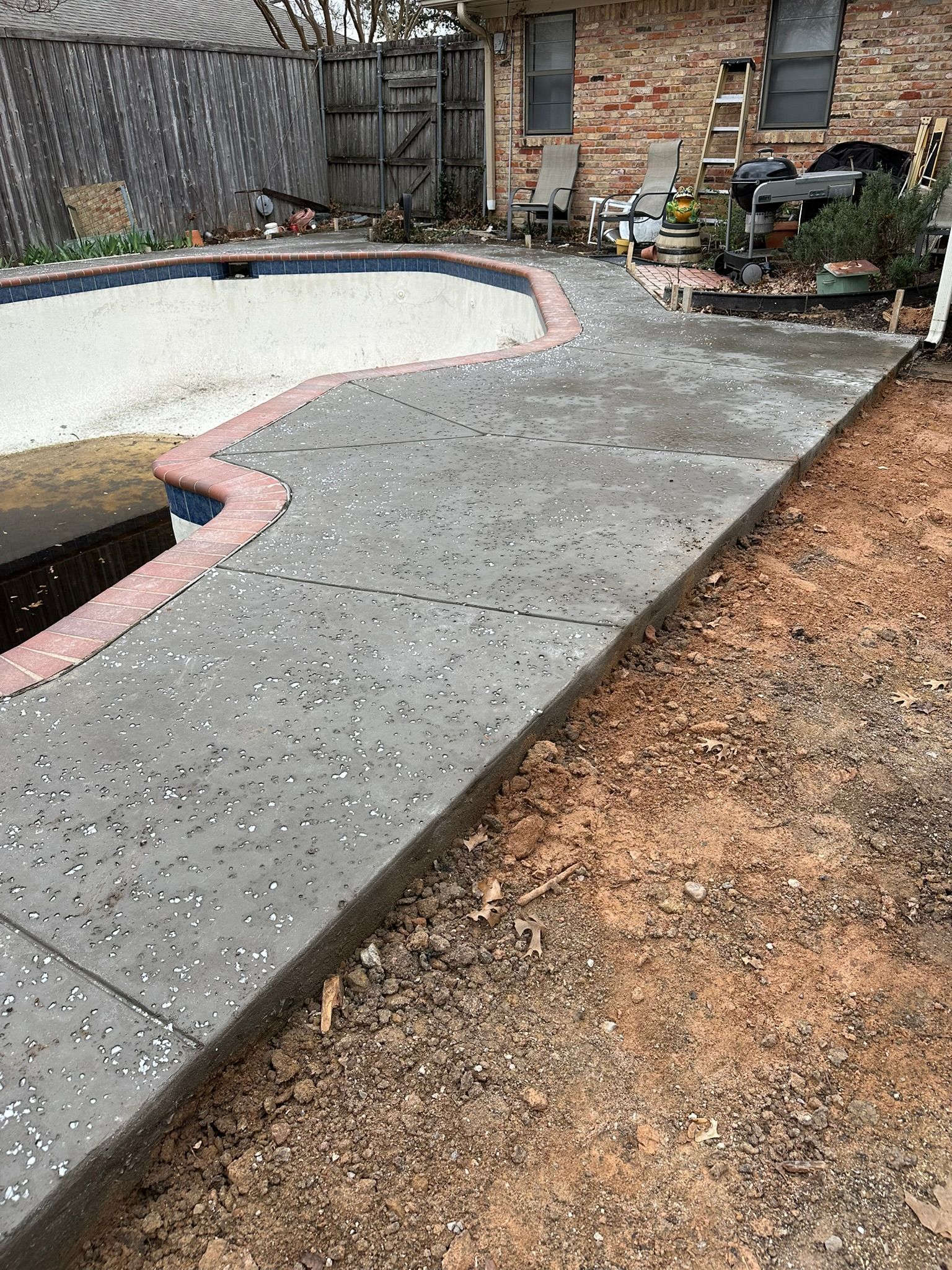 Concrete walkway beside a pool, adjacent to dirt ground. Pool has red brick border.