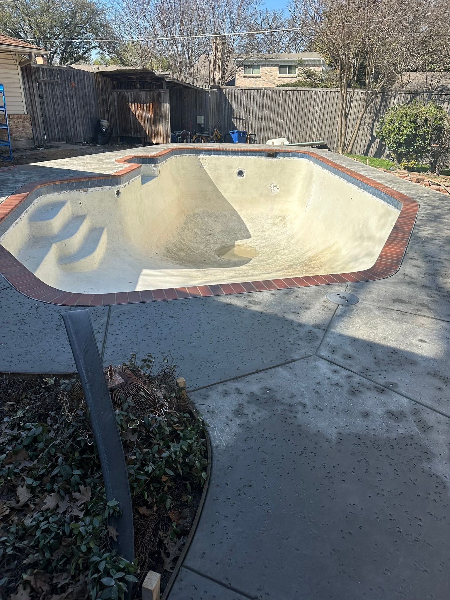 Empty swimming pool with brick coping, surrounded by concrete patio and a wooden fence.