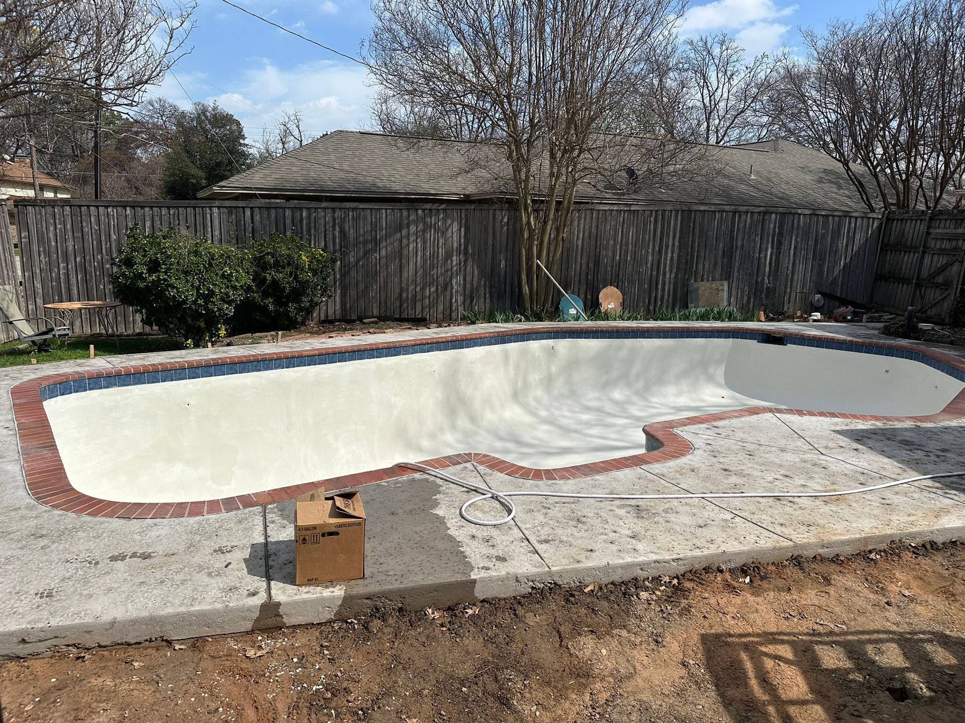 Empty, light-colored swimming pool with red brick border, in backyard with fence and trees.