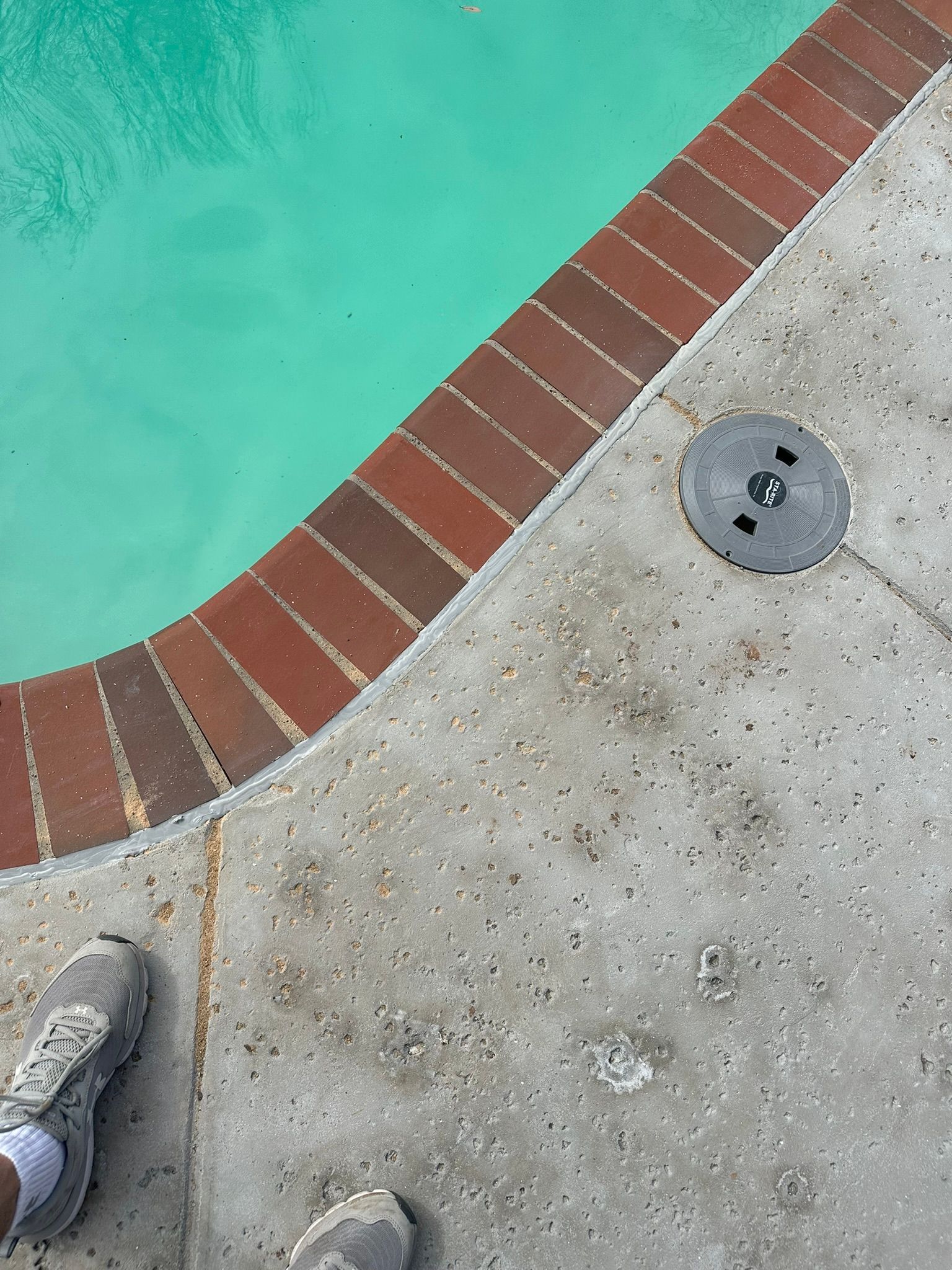 Poolside view: brick edge, turquoise water, concrete surface, a black drain cover, and a person's shoe.
