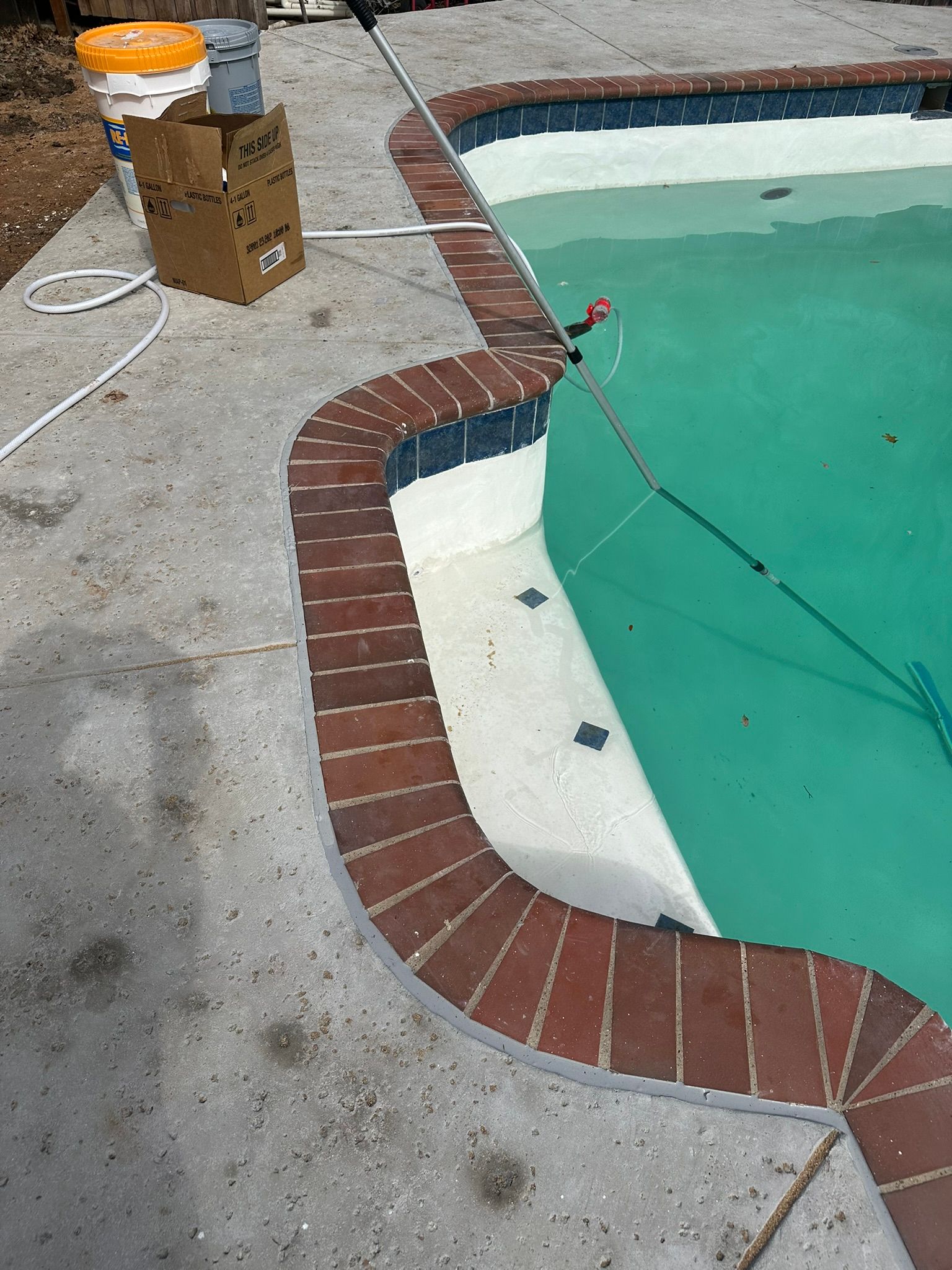 Pool with a red brick coping, teal water, and concrete deck. Equipment and a cardboard box are nearby.