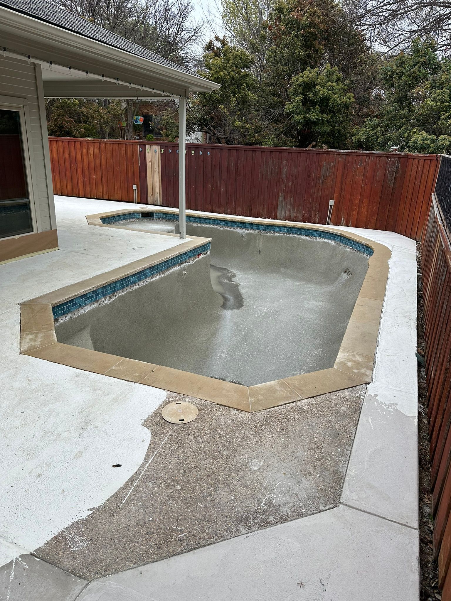 Empty rectangular pool surrounded by concrete and gravel, next to a house and a wooden fence.