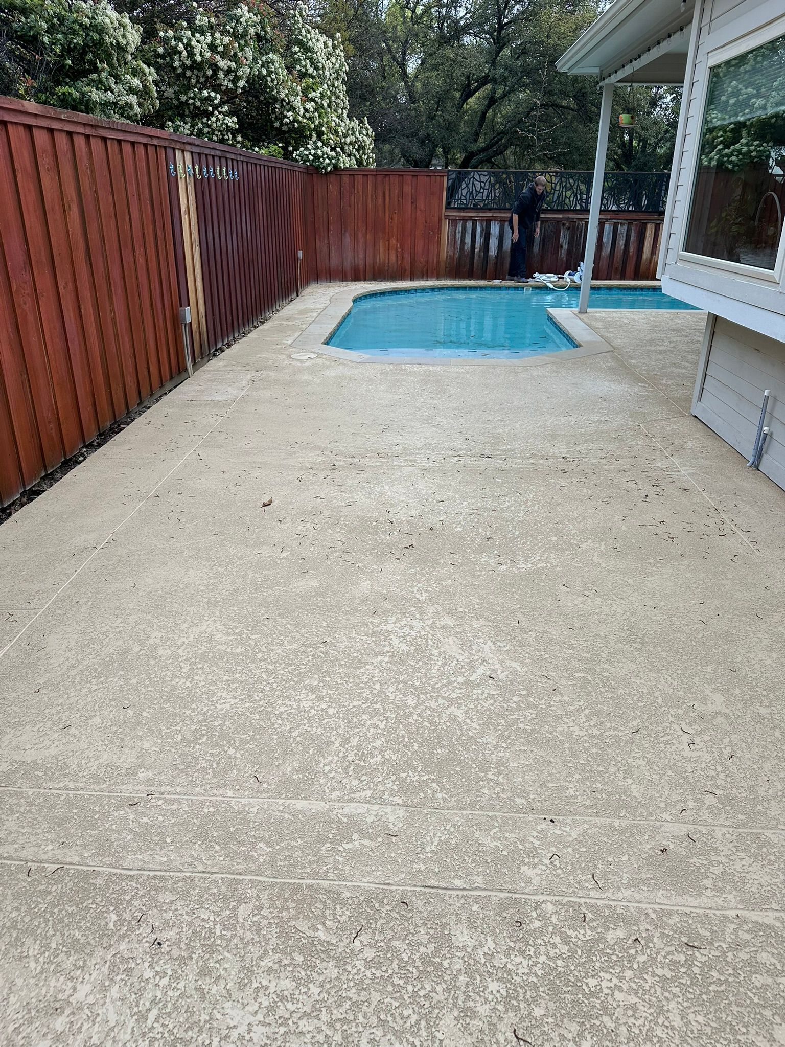 Concrete backyard with small pool, brown fence, and white house.