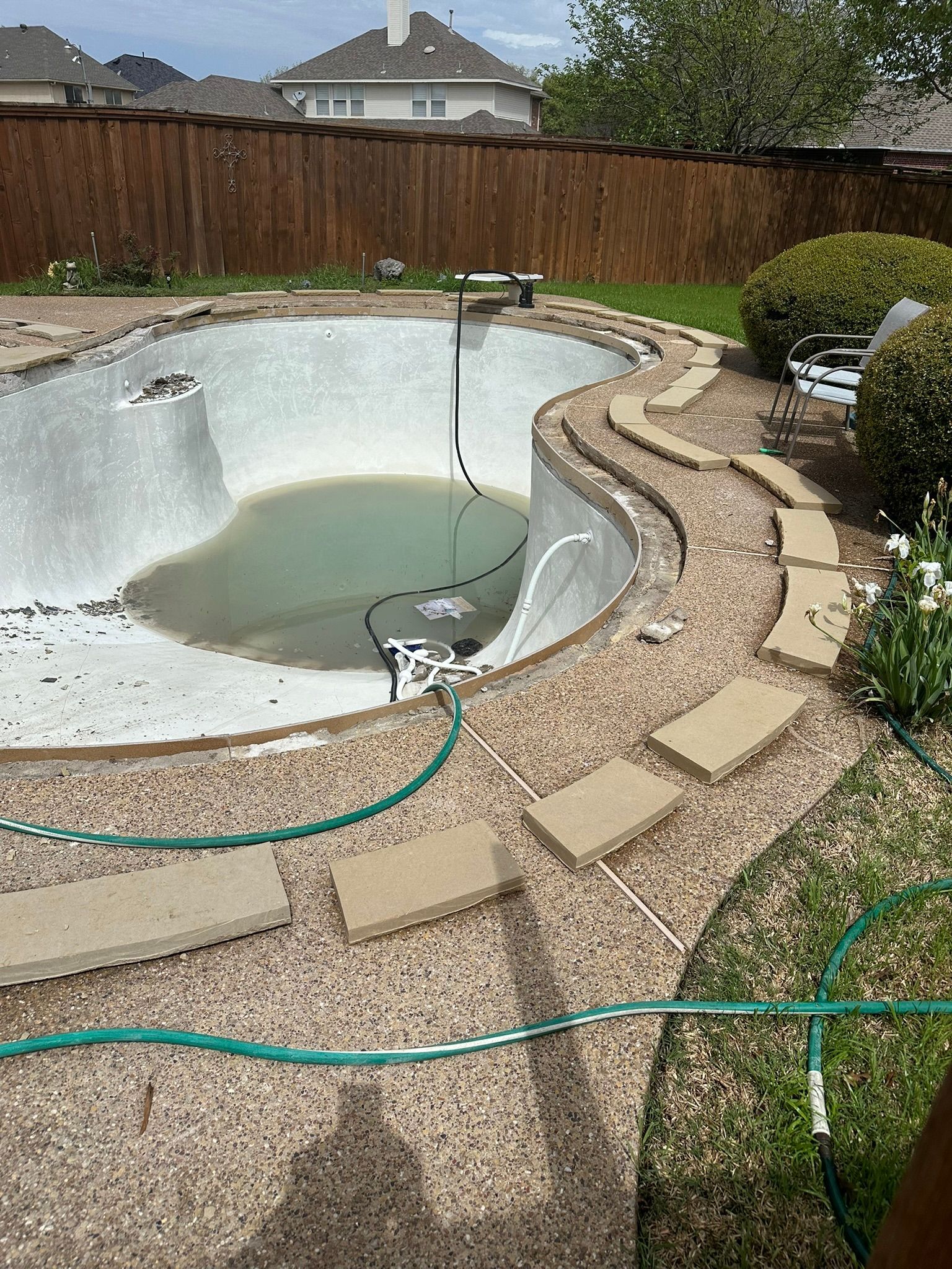 An empty, kidney-shaped swimming pool with green water, surrounded by gravel, stepping stones, and a wooden fence.