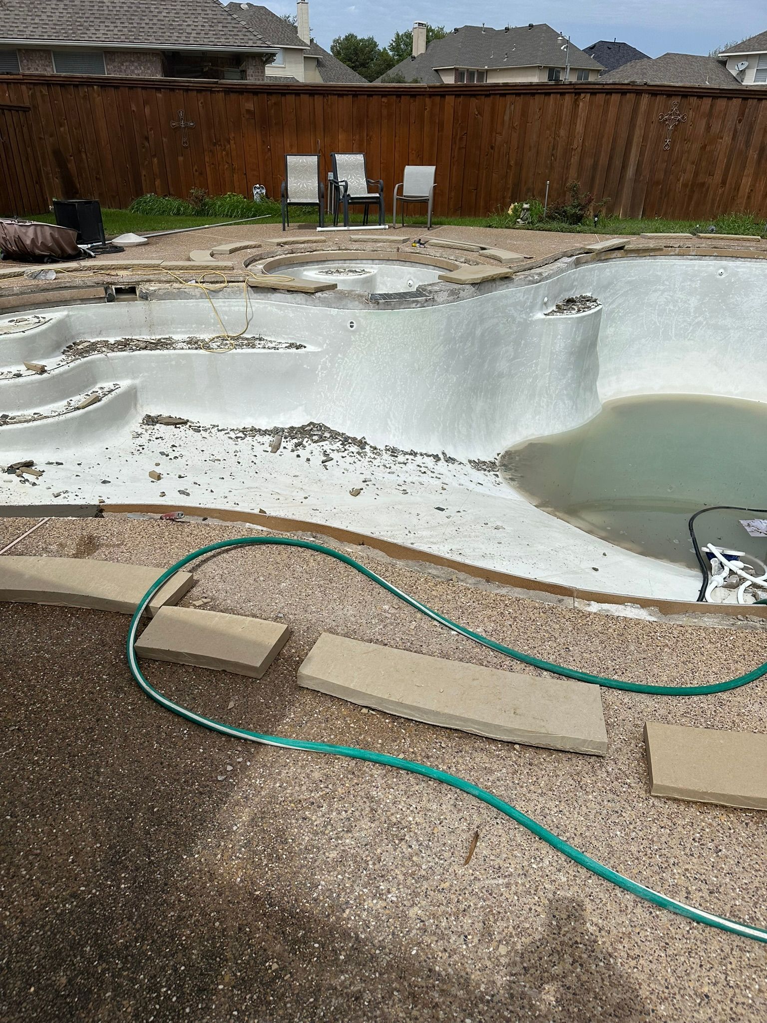 Pool under construction with exposed interior. A green hose lies on the gravel pathway. Brown fence in background.