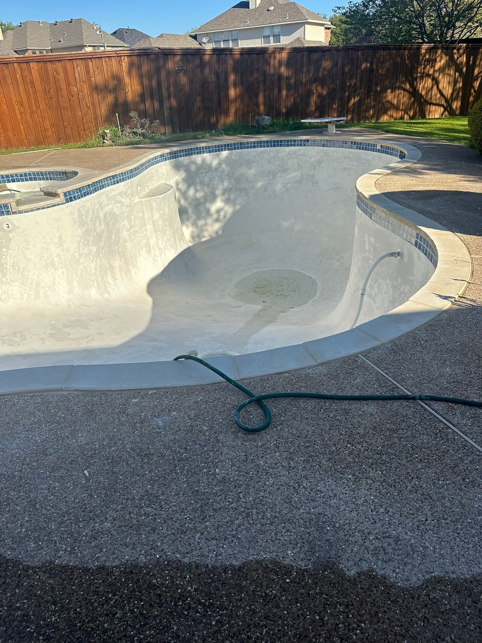 Empty swimming pool with blue tile border, surrounded by concrete and a wooden fence.