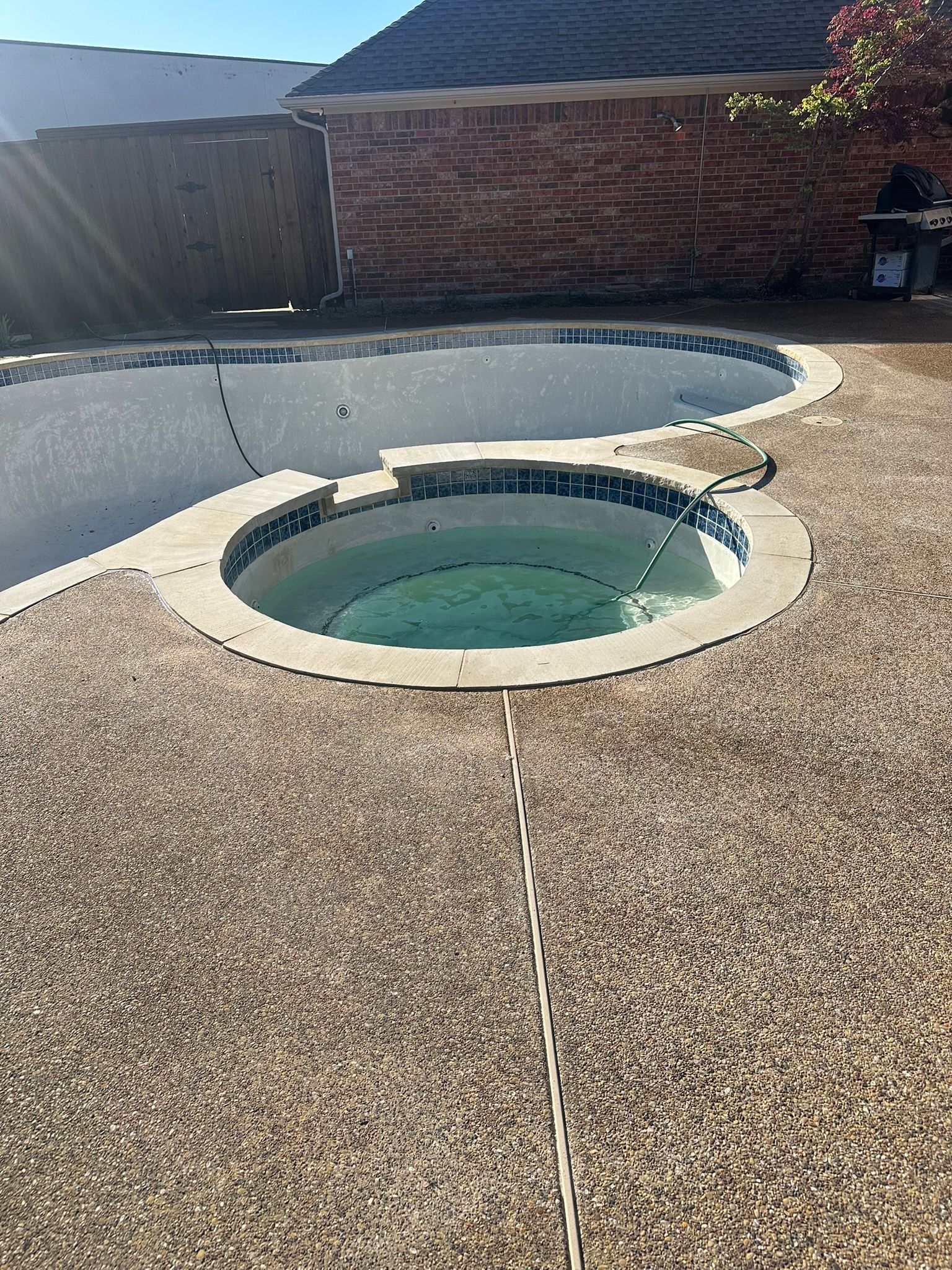 Pool and spa with tan concrete patio, surrounded by brick building and wood fence.