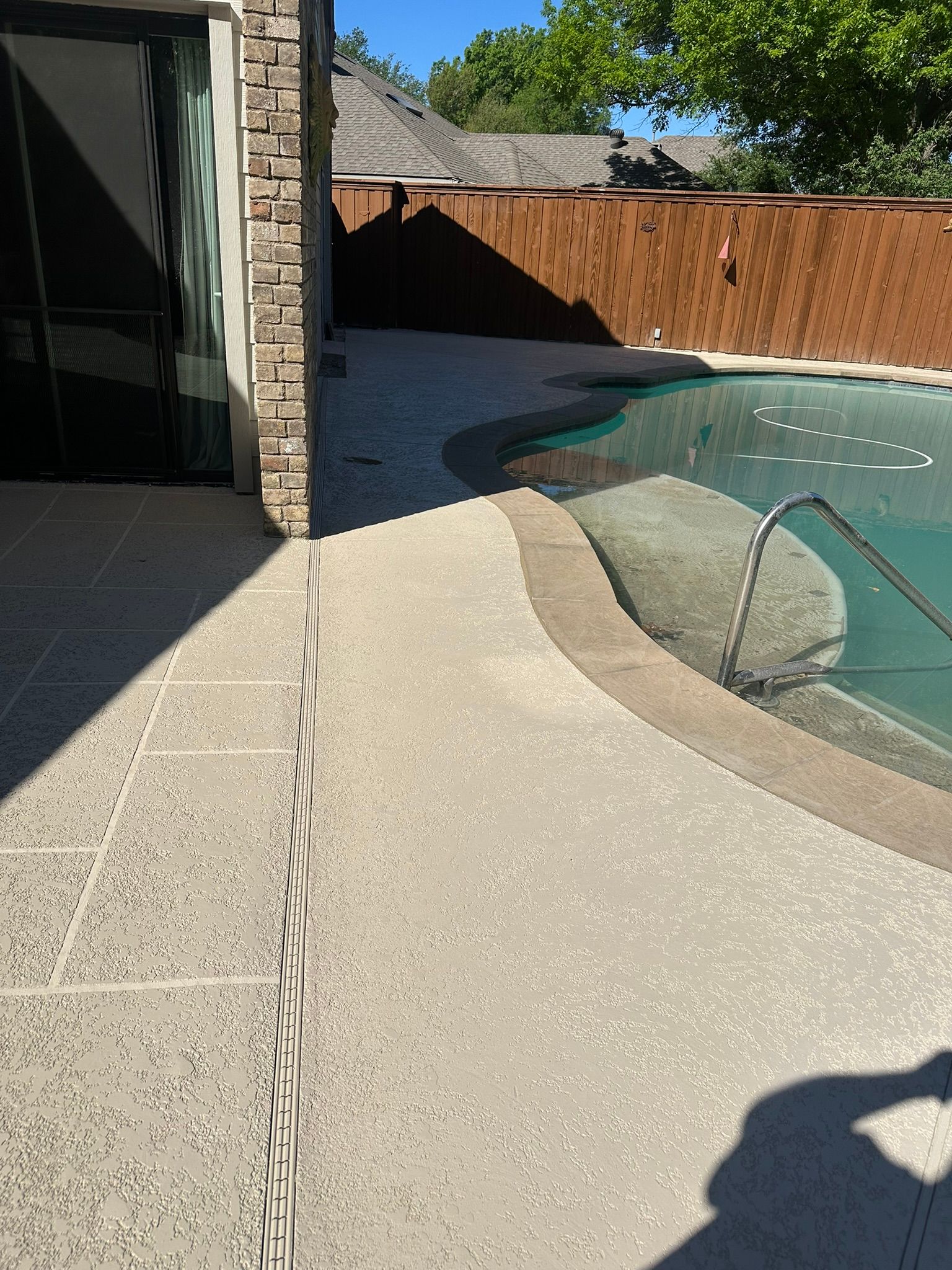Poolside concrete patio with access to a swimming pool, adjacent to a stone wall and wooden fence under a sunny sky.