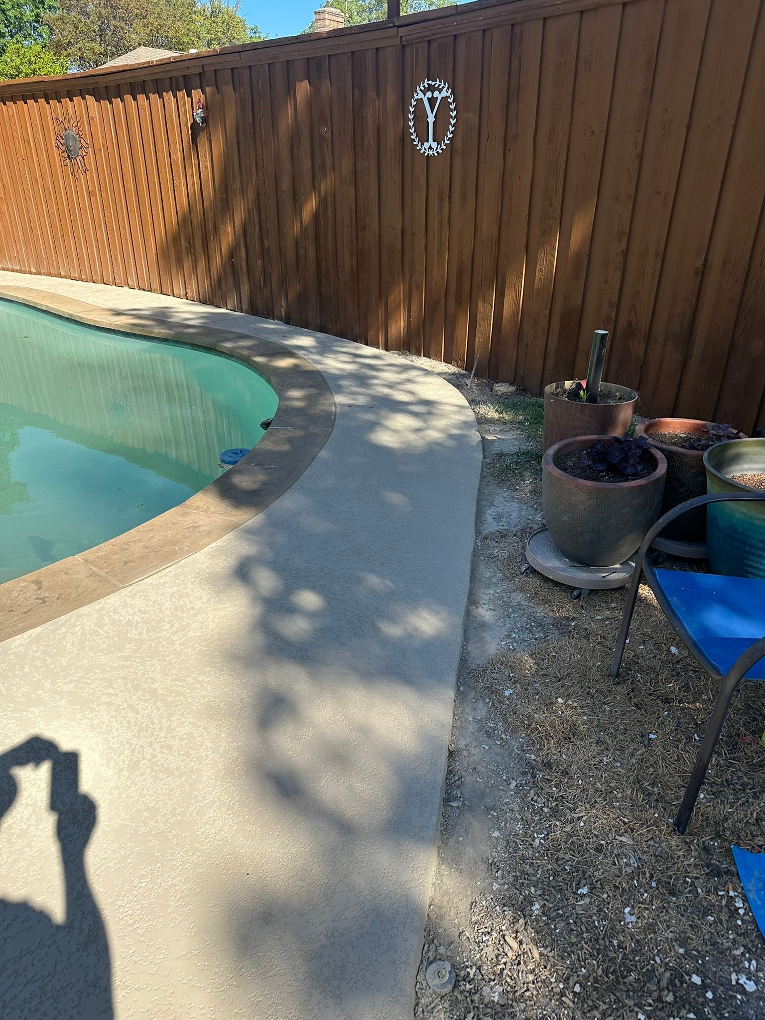 Poolside view with concrete border, wooden fence, potted plants, and gravel ground.