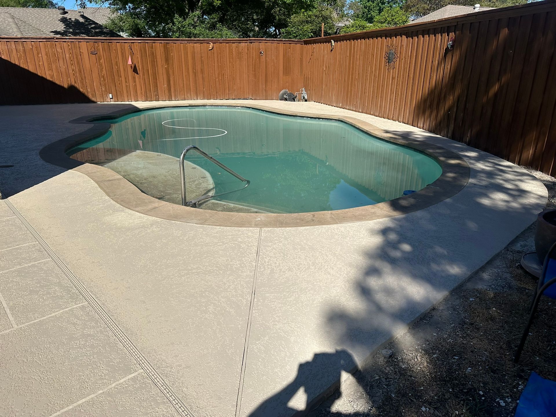 Swimming pool surrounded by concrete, wooden fence in the background, sunny day.