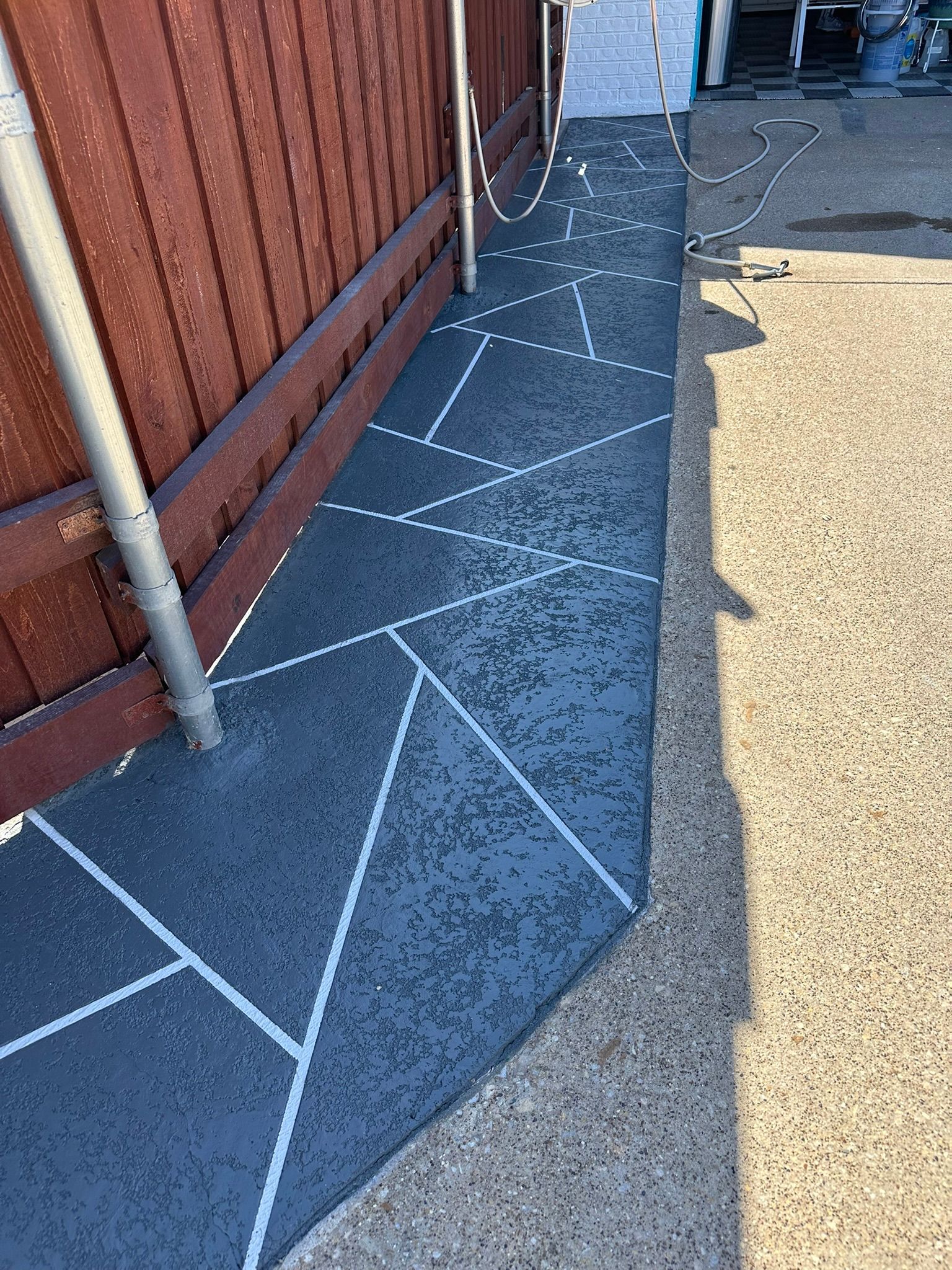 Concrete walkway with a geometric pattern, next to a brown wooden fence and a paved surface.
