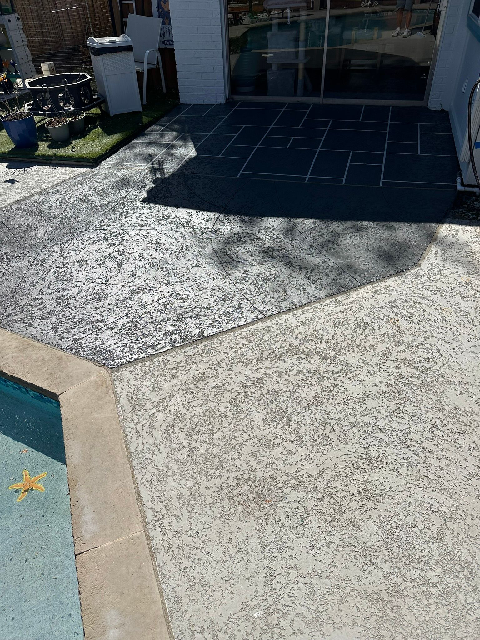 Concrete patio with a pool and a dark geometric patterned section near a glass door.