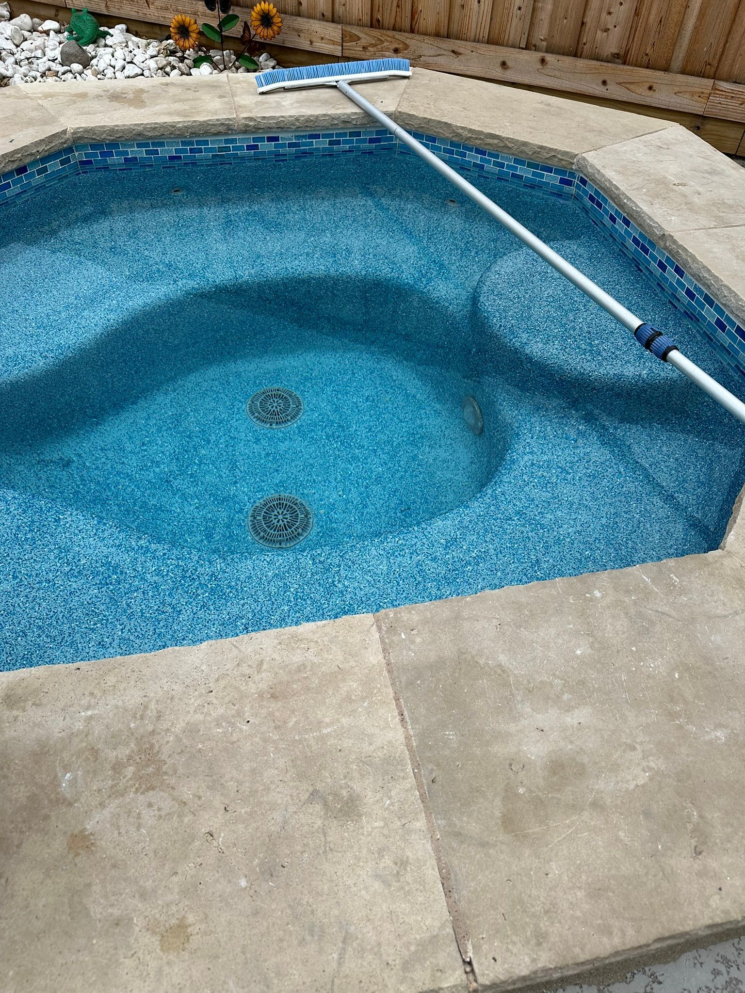 Small, blue-tiled pool being cleaned with a pool brush. Concrete patio surrounds the pool.