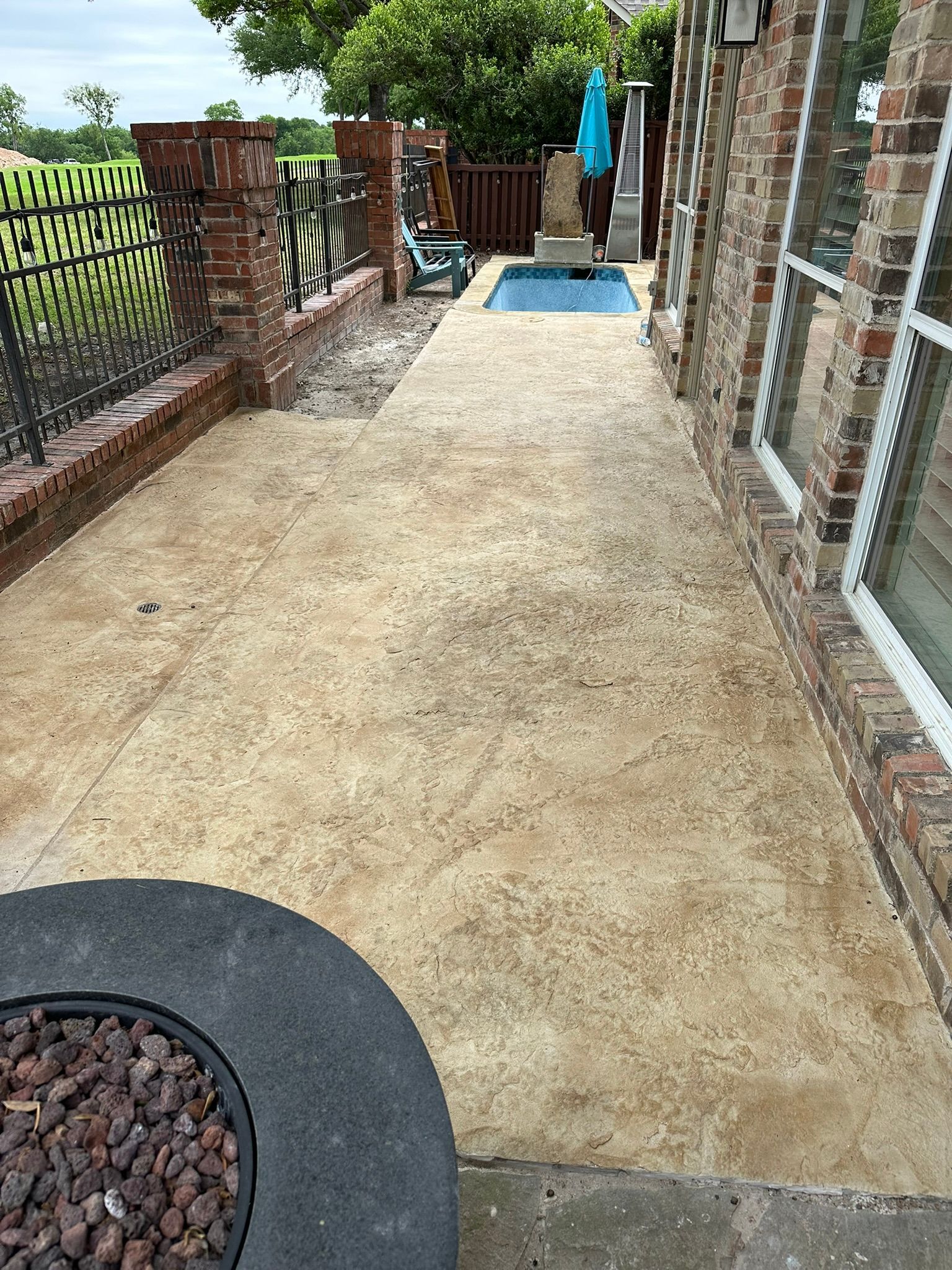 Concrete patio next to a brick house and decorative fence, with a small pool in the distance.