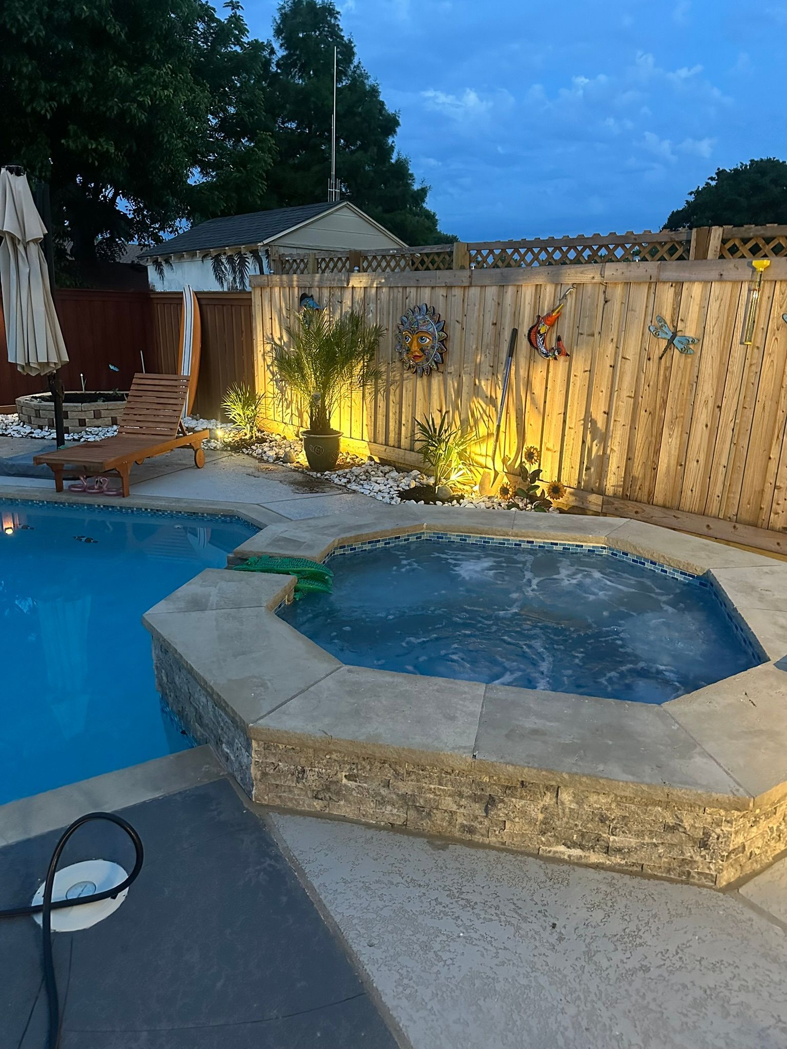 Pool and hot tub at dusk, surrounded by wooden fence with outdoor lighting, plants, and a lounge chair.