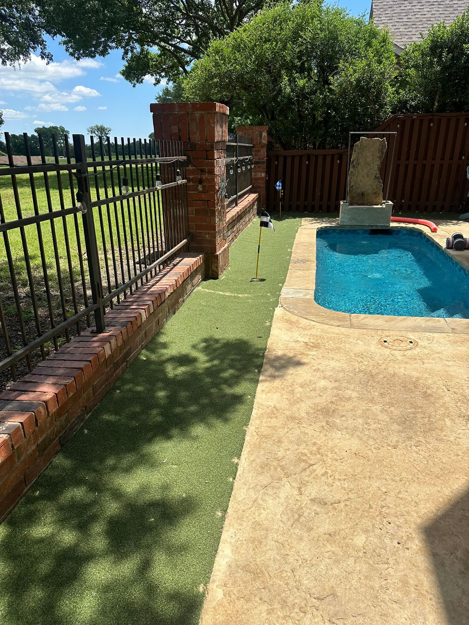 Backyard with a mini golf green, pool, and brick pillars next to a wrought-iron fence.