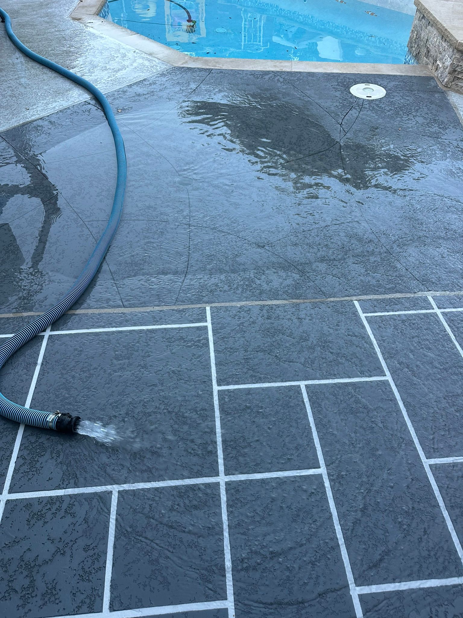 Water sprays from a blue hose onto wet, dark gray stone pavers surrounding a swimming pool.
