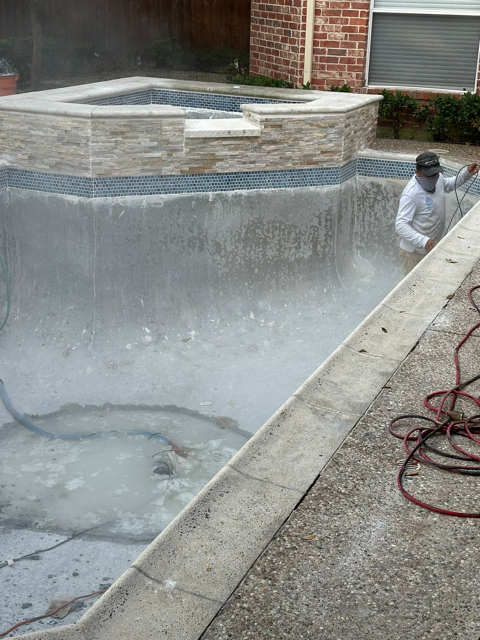 Man wearing protective gear power washing a swimming pool, splashing water.