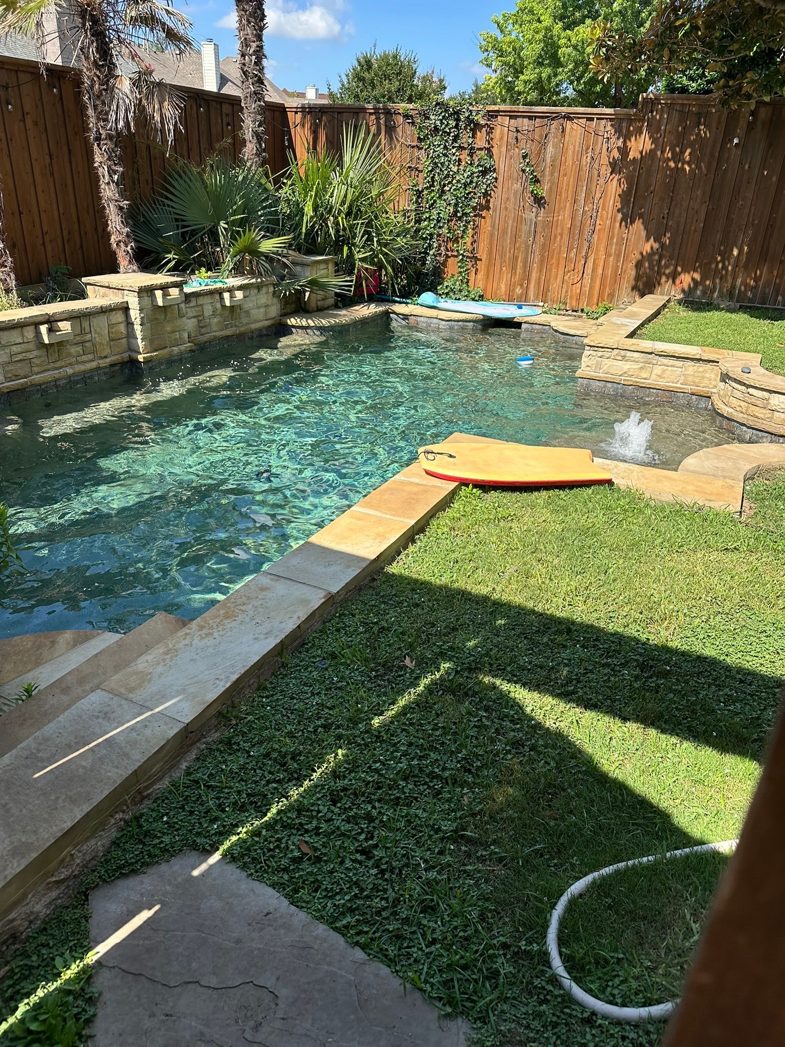Pool with clear water, surrounded by stone and greenery, wooden fence in the background.