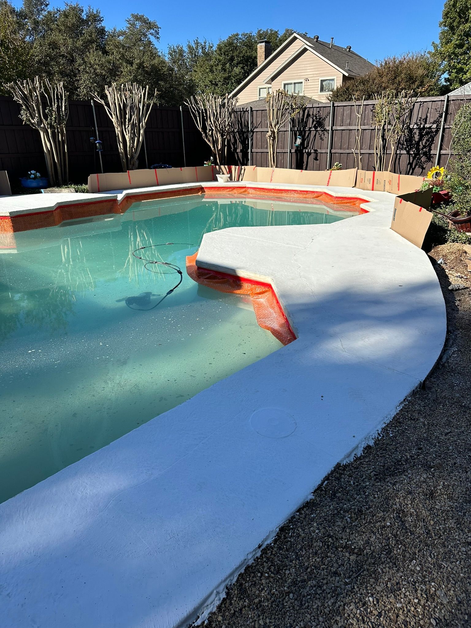 Pool with white concrete border, turquoise water, and a fence and house in the background. Sunny day.