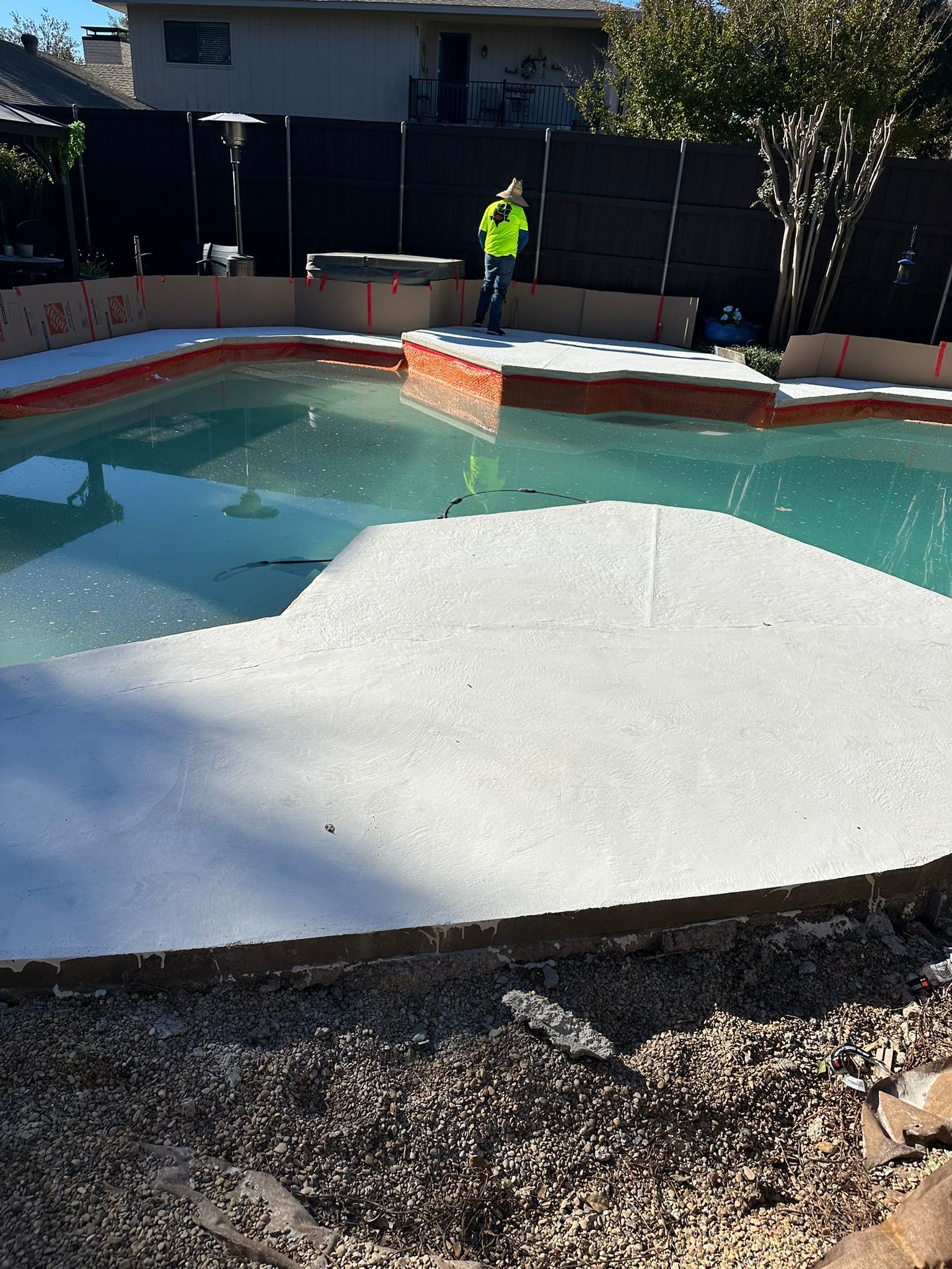 Pool construction: worker on a deck, water in pool, light concrete patio, gravel border.